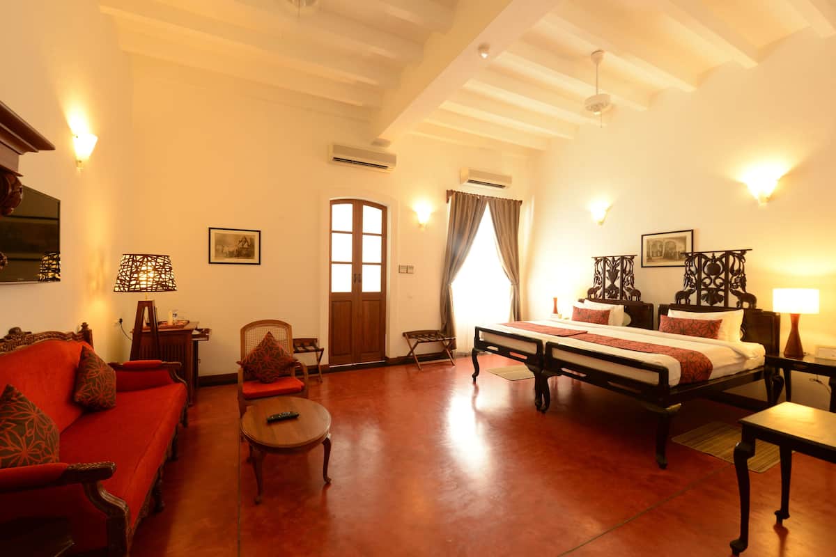 Une chambre d’hôtel avec un lit, un canapé, une petite table et une chaise. Il y a des lampes fixées au mur et une porte avec une fenêtre.