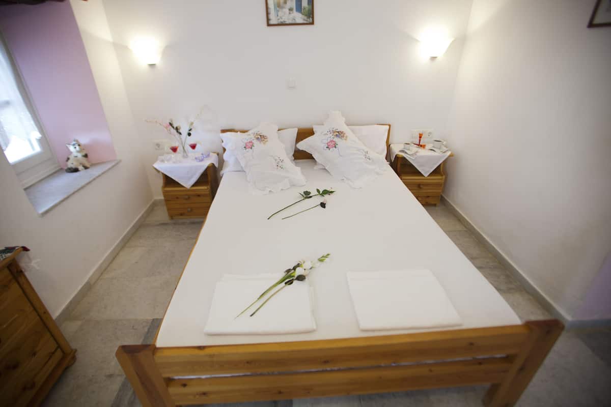 Ein ordentlich bezogenes Bett mit weißen Leinen und floralen Verzierungen, ein Nachttisch aus Holz mit einer Lampe und ein kleines Fenster mit einer Katze auf dem Sims.