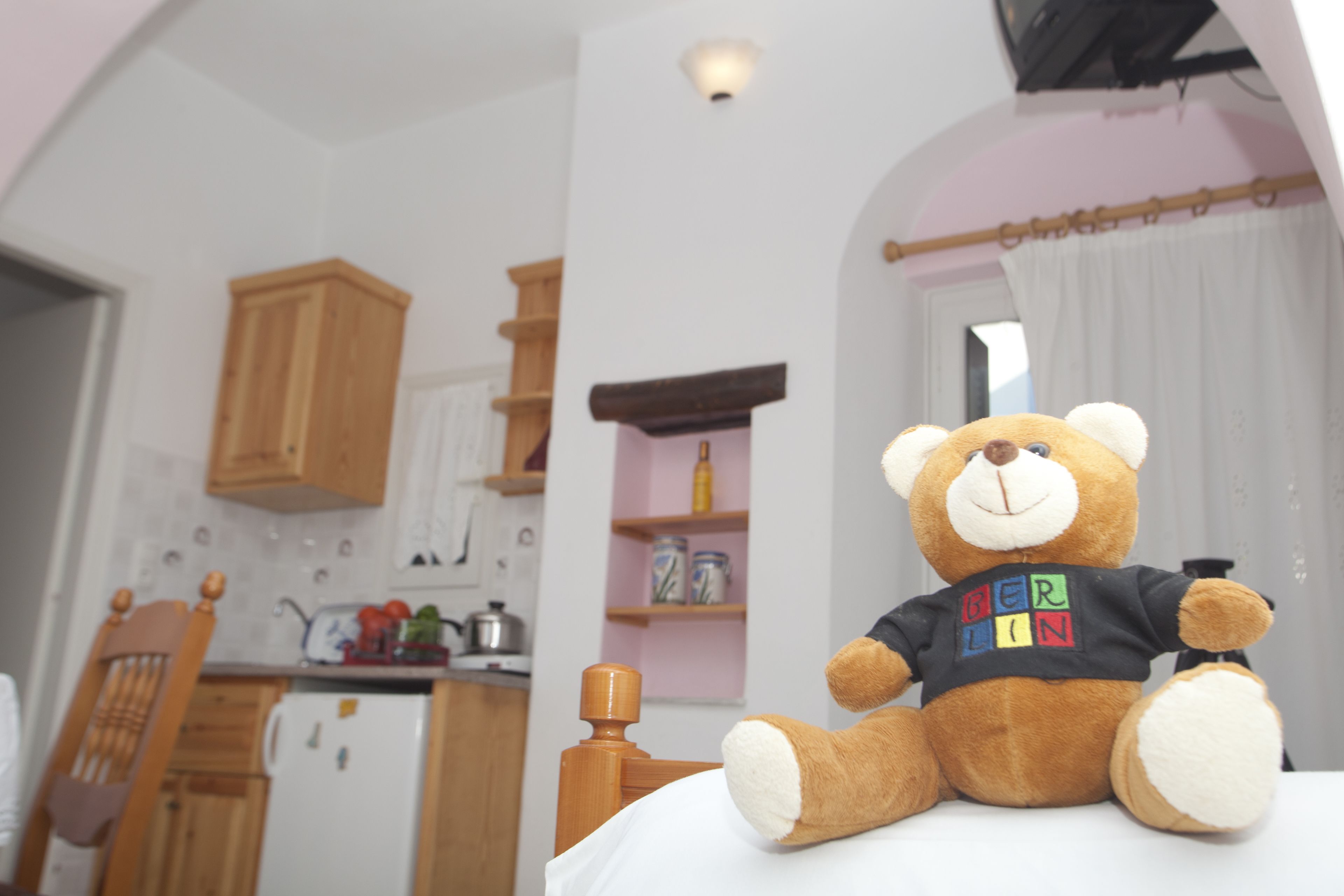 Un ours en peluche est assis sur un lit dans une pièce où l’on aperçoit une cuisine au fond.