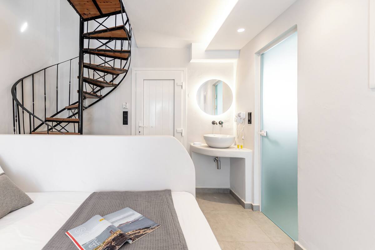 Ein modernes Schlafzimmer mit Wendeltreppe, einem großen Bett, einem Nachttisch mit einer Zeitschrift und einem Badezimmer mit rundem Spiegel und Waschbecken.