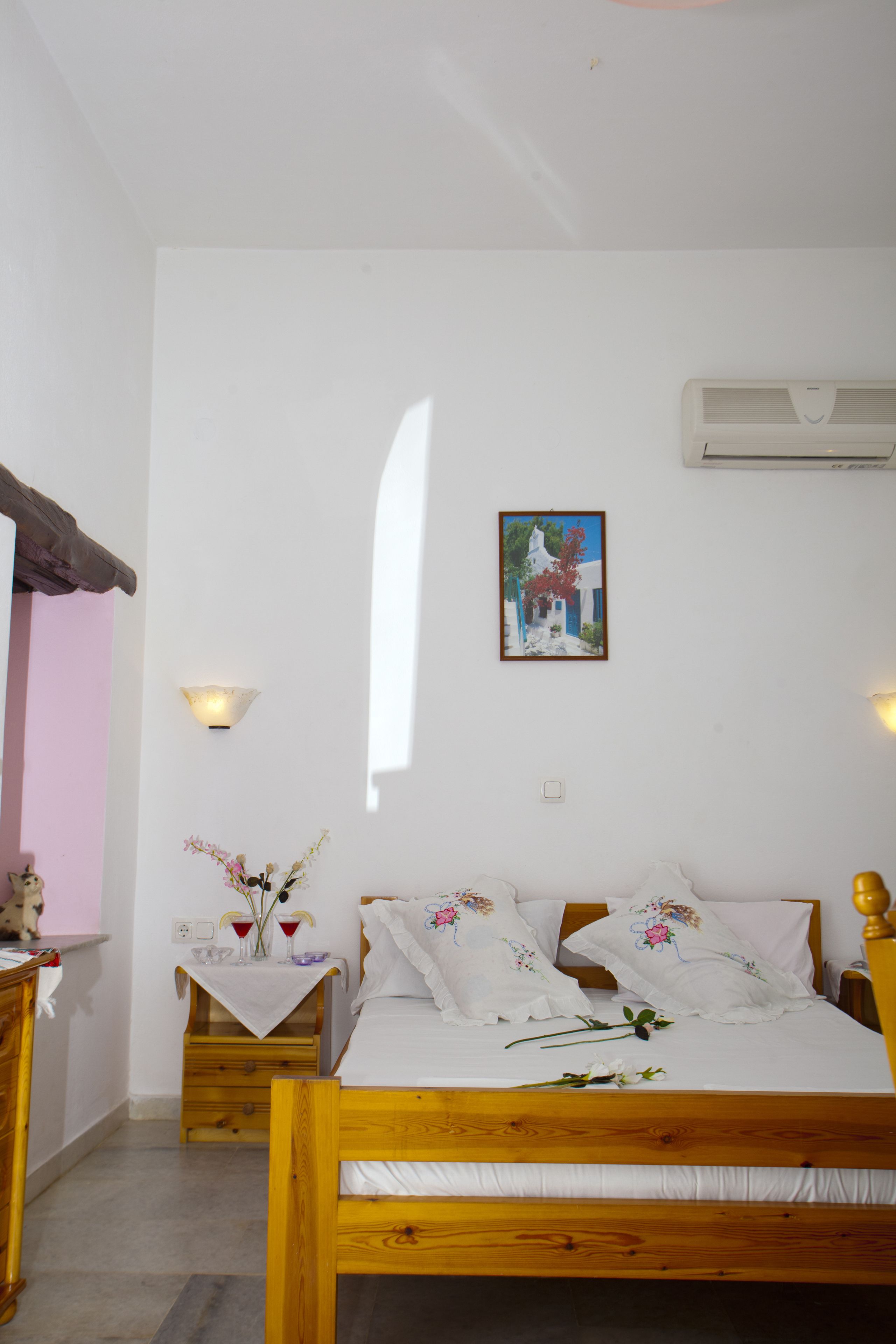 Un dormitorio con cama de madera, ropa de cama blanca, una mesita con un jarrón y copas de vino, y un aire acondicionado instalado en la pared.