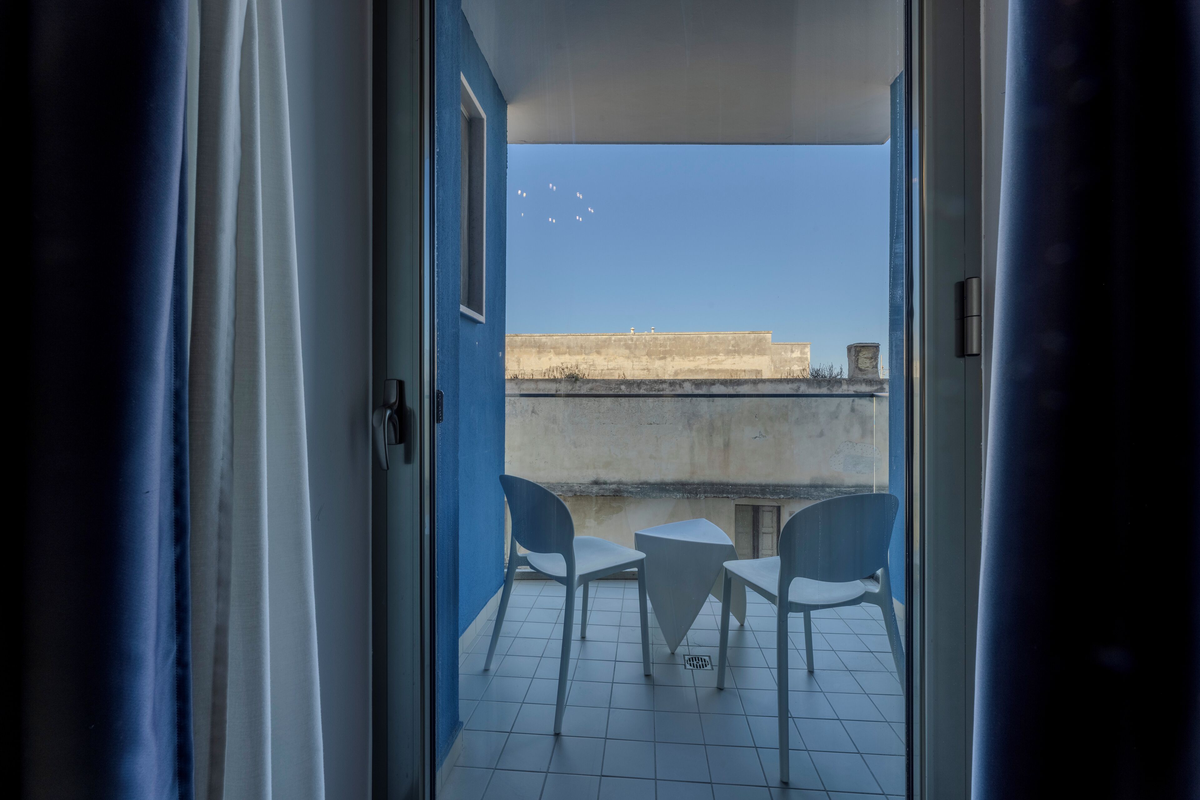 An open door leading to a balcony with two white chairs and a small table, overlooking a building with a clear sky.