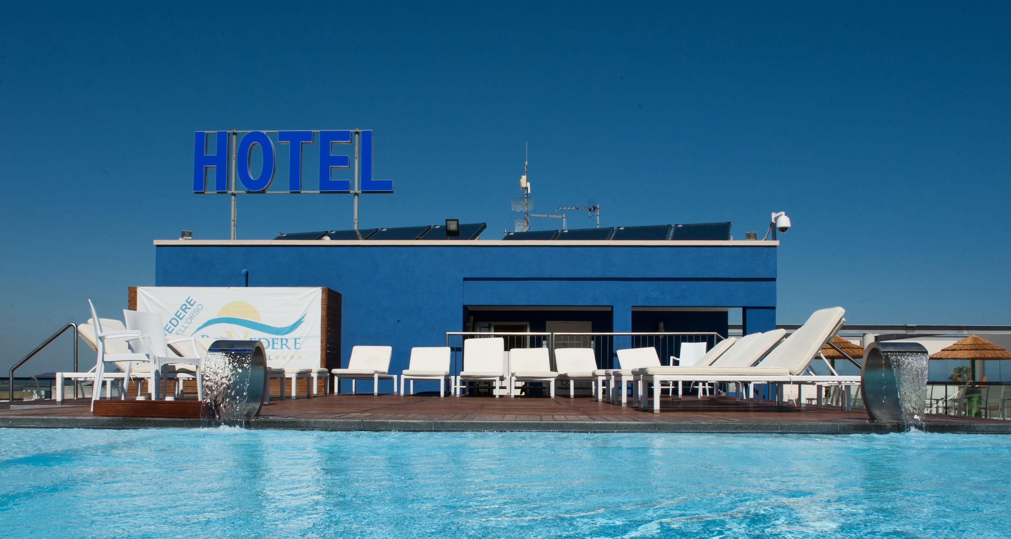A hotel with a large blue sign, a pool with white lounge chairs, and a water feature.