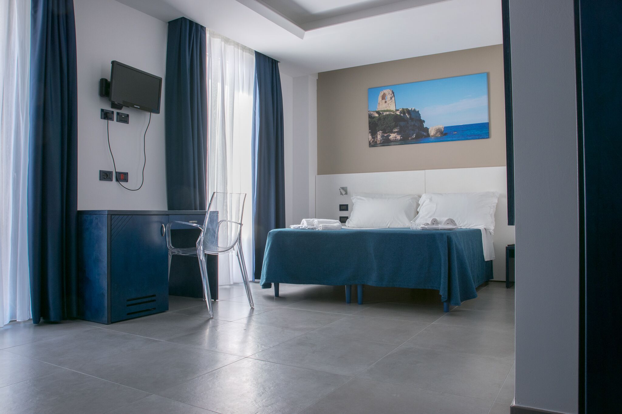 Una camera d'albergo con un letto, una scrivania, una sedia, una TV a parete e un quadro raffigurante una scena costiera.