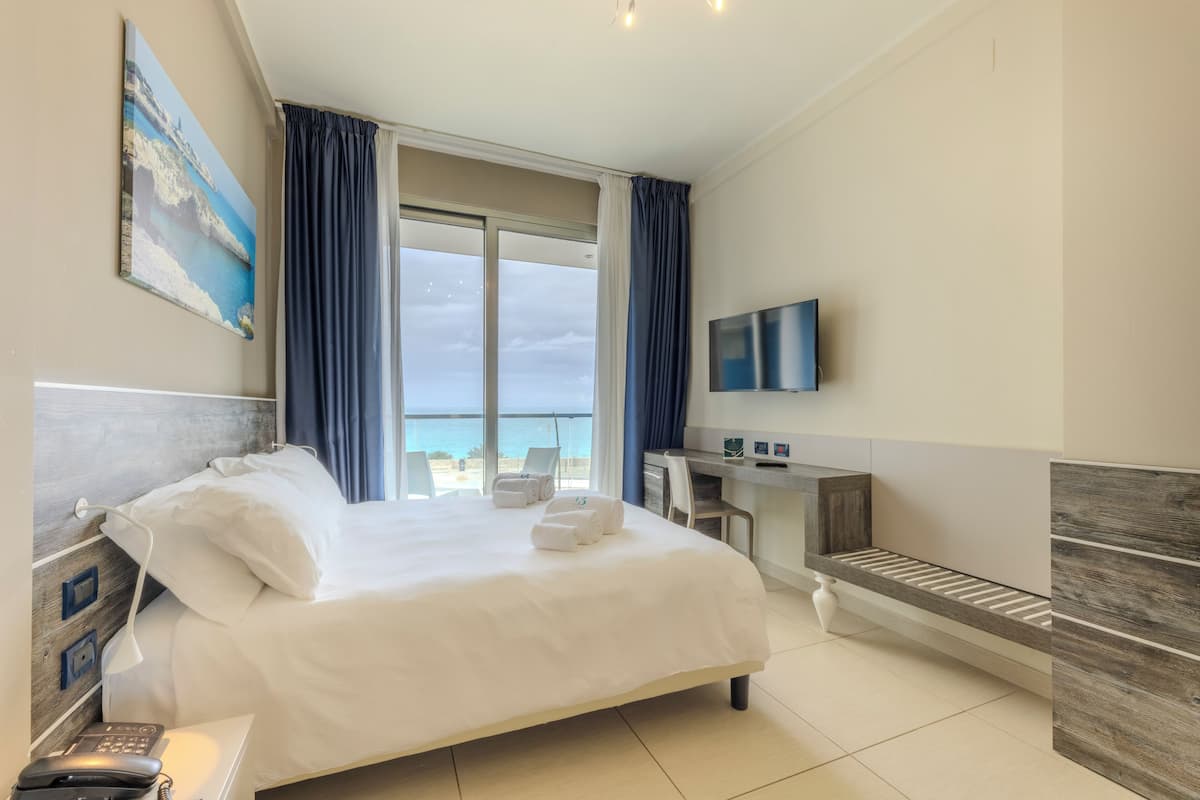 A hotel room with a bed, a desk, a TV, and a view of the sea.