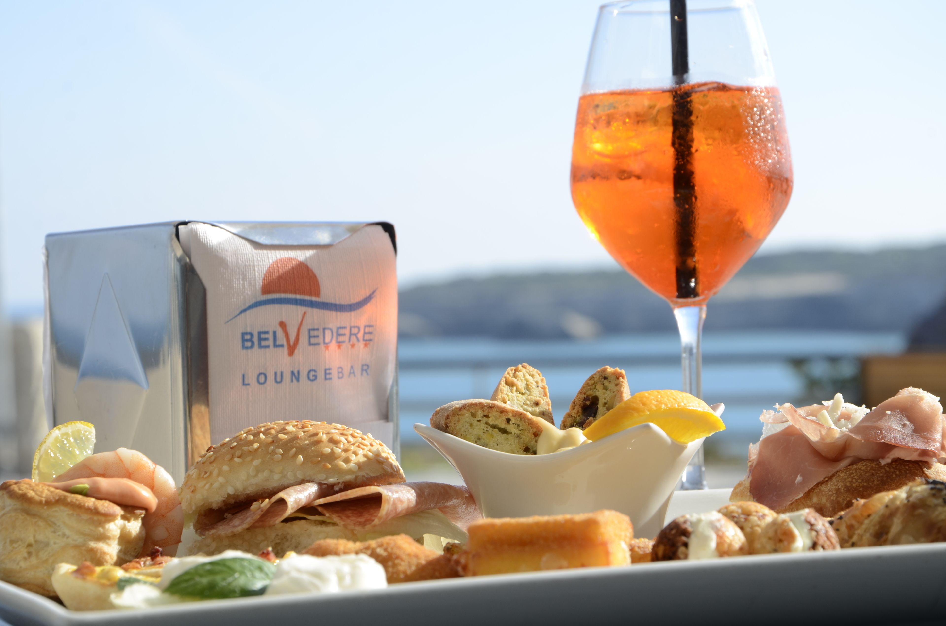 Varie pietanze tra cui panini, pasticcini e un bicchiere di succo d'arancia, con vista sul mare sullo sfondo.