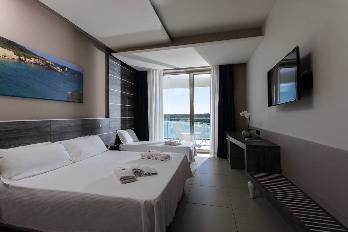 A hotel room with two beds, a TV, and a balcony with a view of the sea.