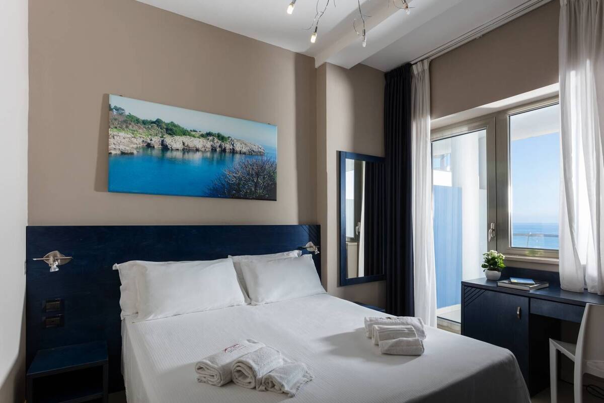 A hotel room with a large bed, a desk, a chair, a window with a view of the sea, and a painting on the wall.