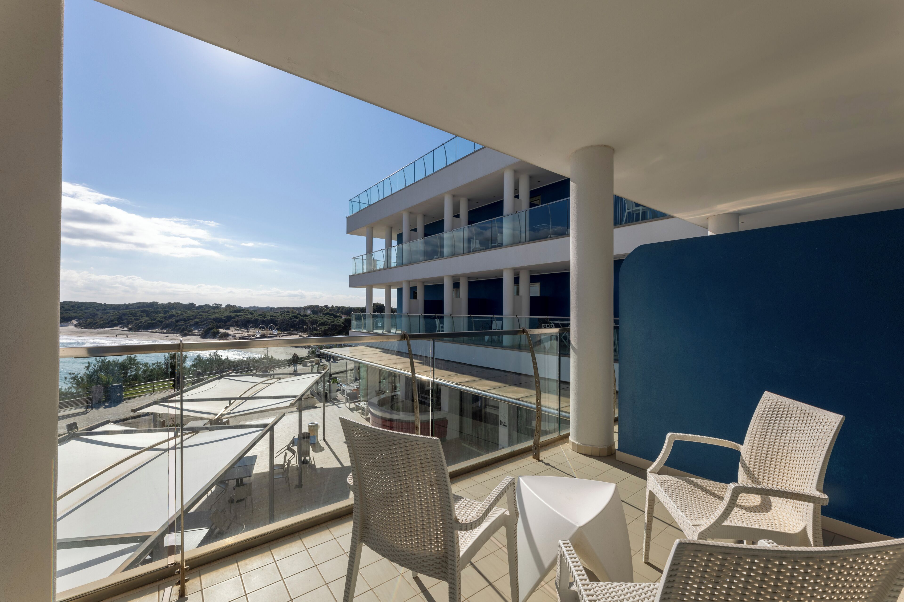 Un balcon doté de chaises et d’une table en osier blanc, offrant une vue sur une plage et un immeuble à multiples balcons.