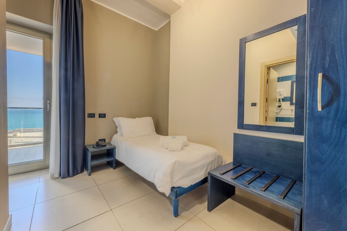 A hotel room with a bed, a bedside table, a bench, a large mirror, and a view of the sea.