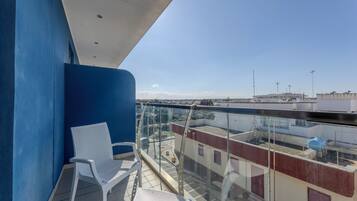 Un balcón con una silla blanca, barandilla de vidrio transparente y vista a edificios distantes.
