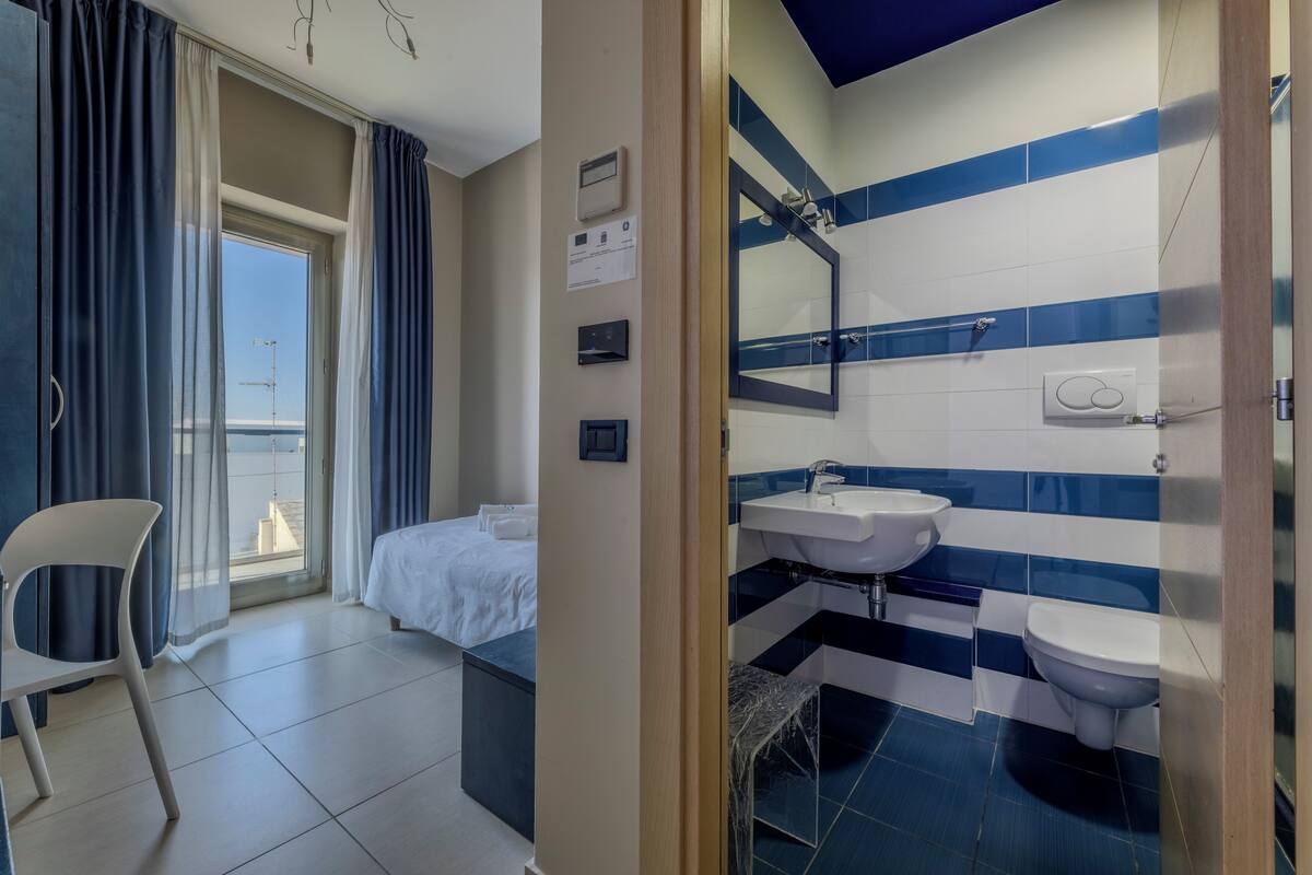 A hotel room with a bed, a chair, a bathroom with a sink and toilet, and a view of the city through the window.