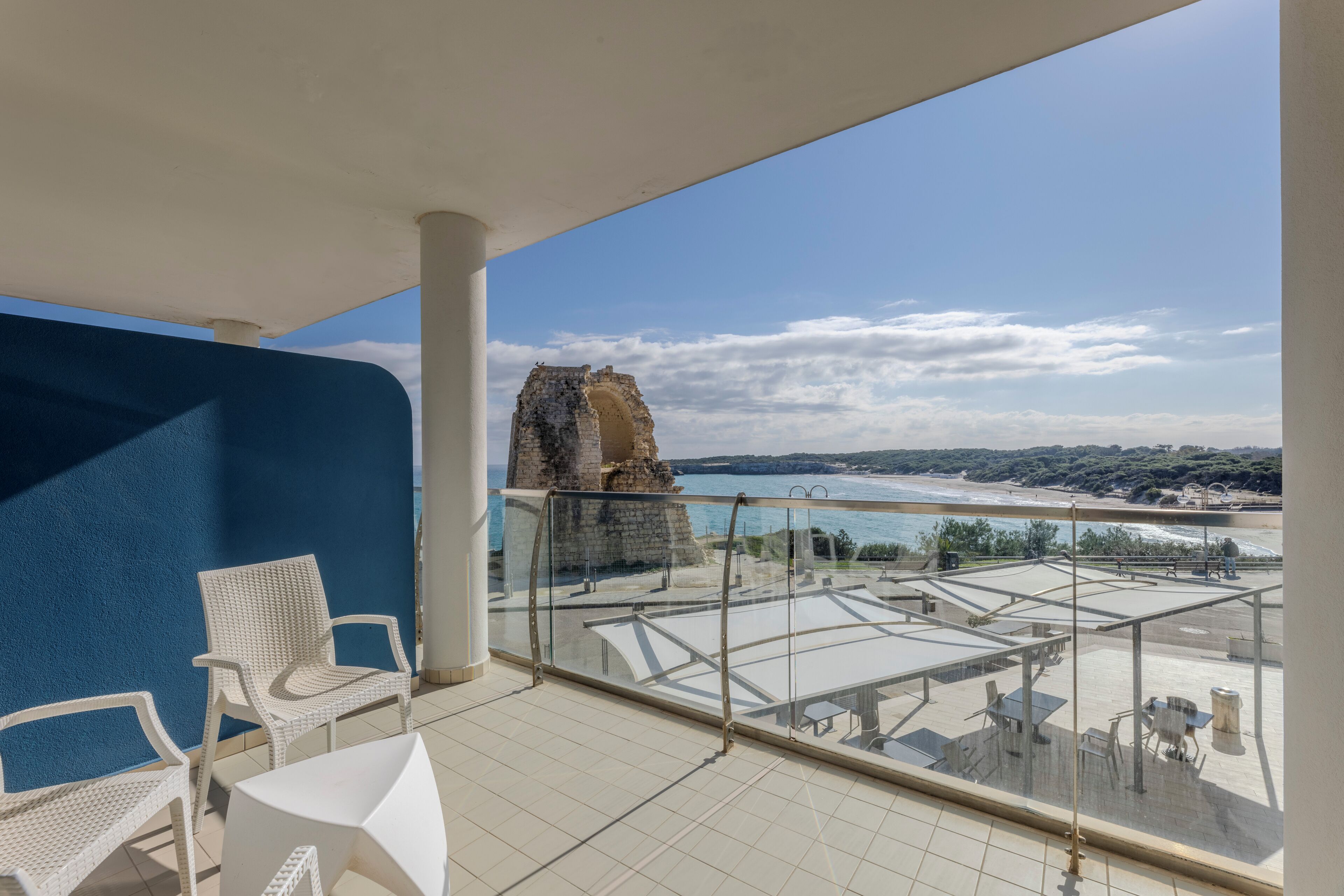 Un balcon avec des chaises et des tables blanches, donnant sur une zone côtière avec une structure en pierre et une plage.
