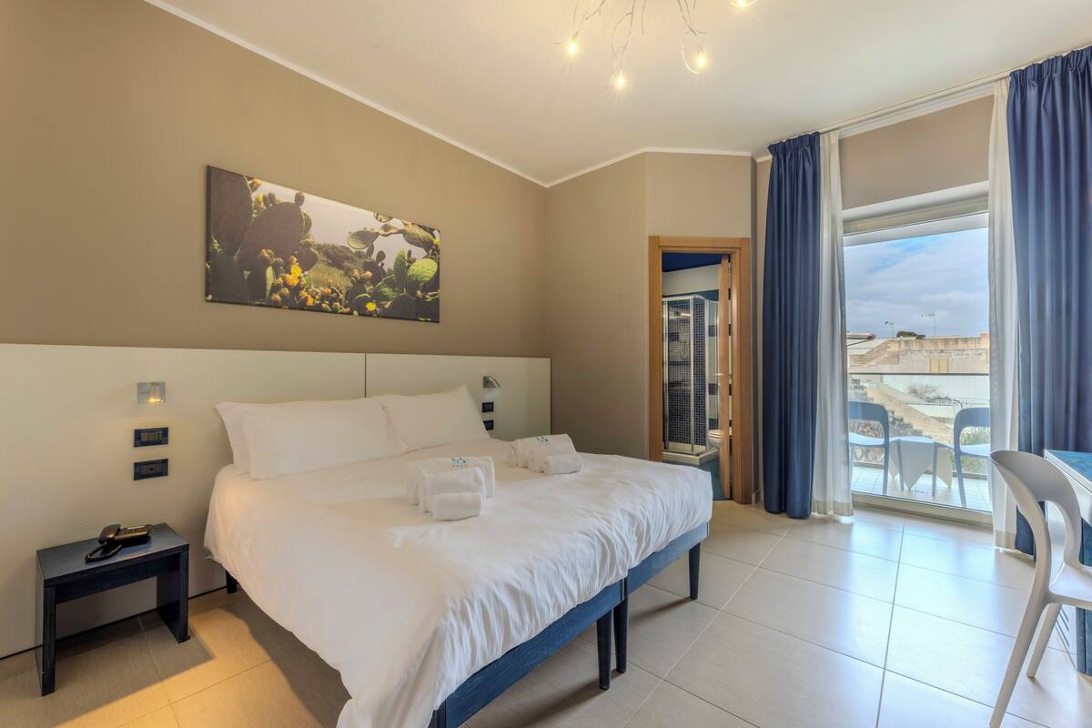 A hotel room with a large bed, a bedside table with a phone, a wall-mounted light, a painting, and a balcony with a chair.