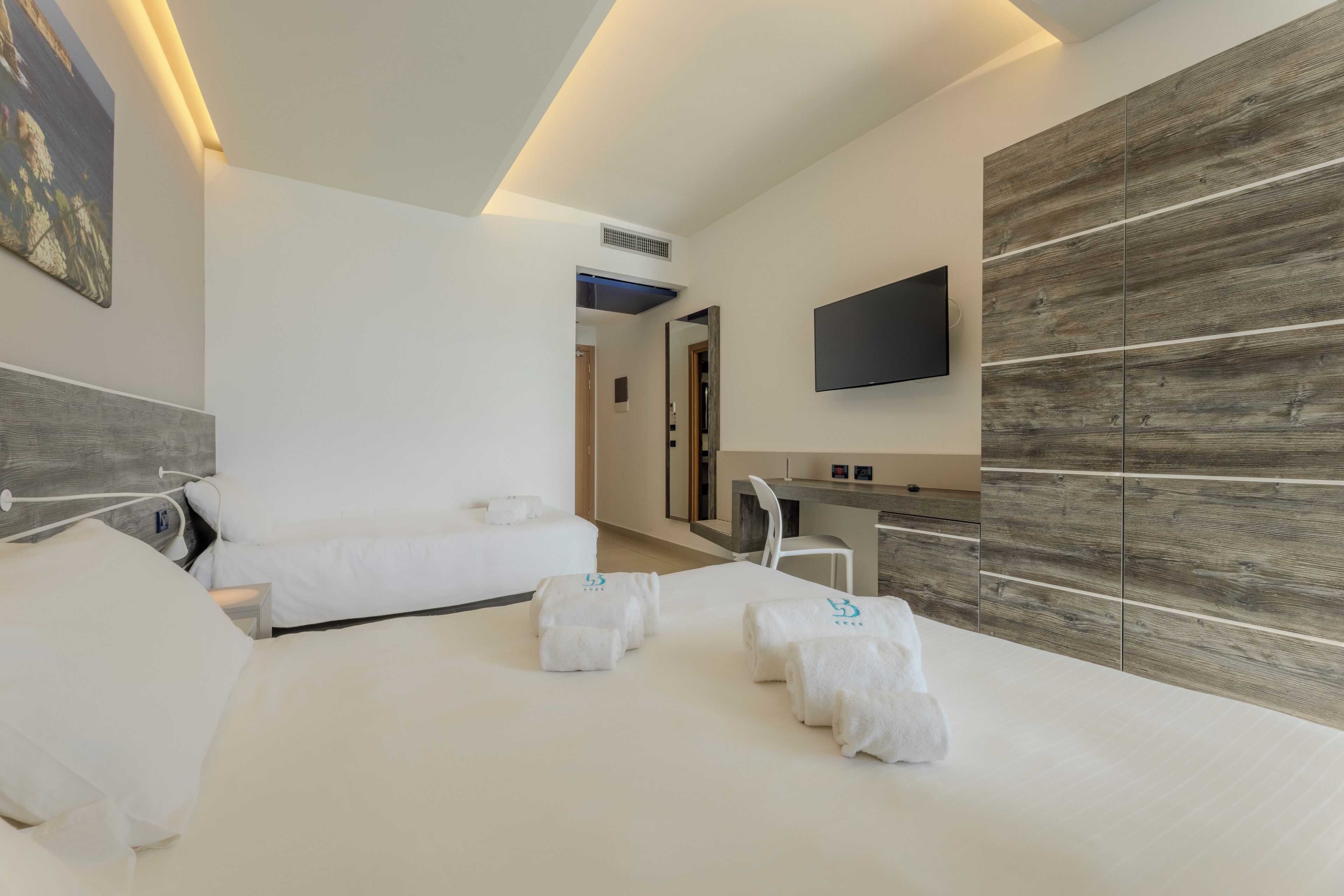 Ein modernes Hotelzimmer mit einem großen Bett, einem Einzelbett, einem Schreibtisch und einem an der Wand befestigten Fernseher.