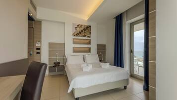 A modern hotel room with a large bed, a desk, and a view of the sea.
