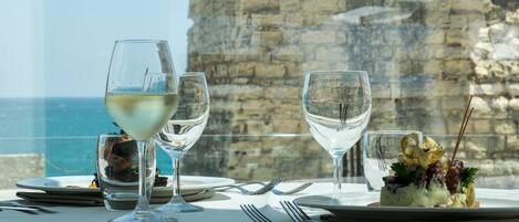 A table set for dining with two wine glasses, a dessert plate, and a view of the sea through a stone wall.