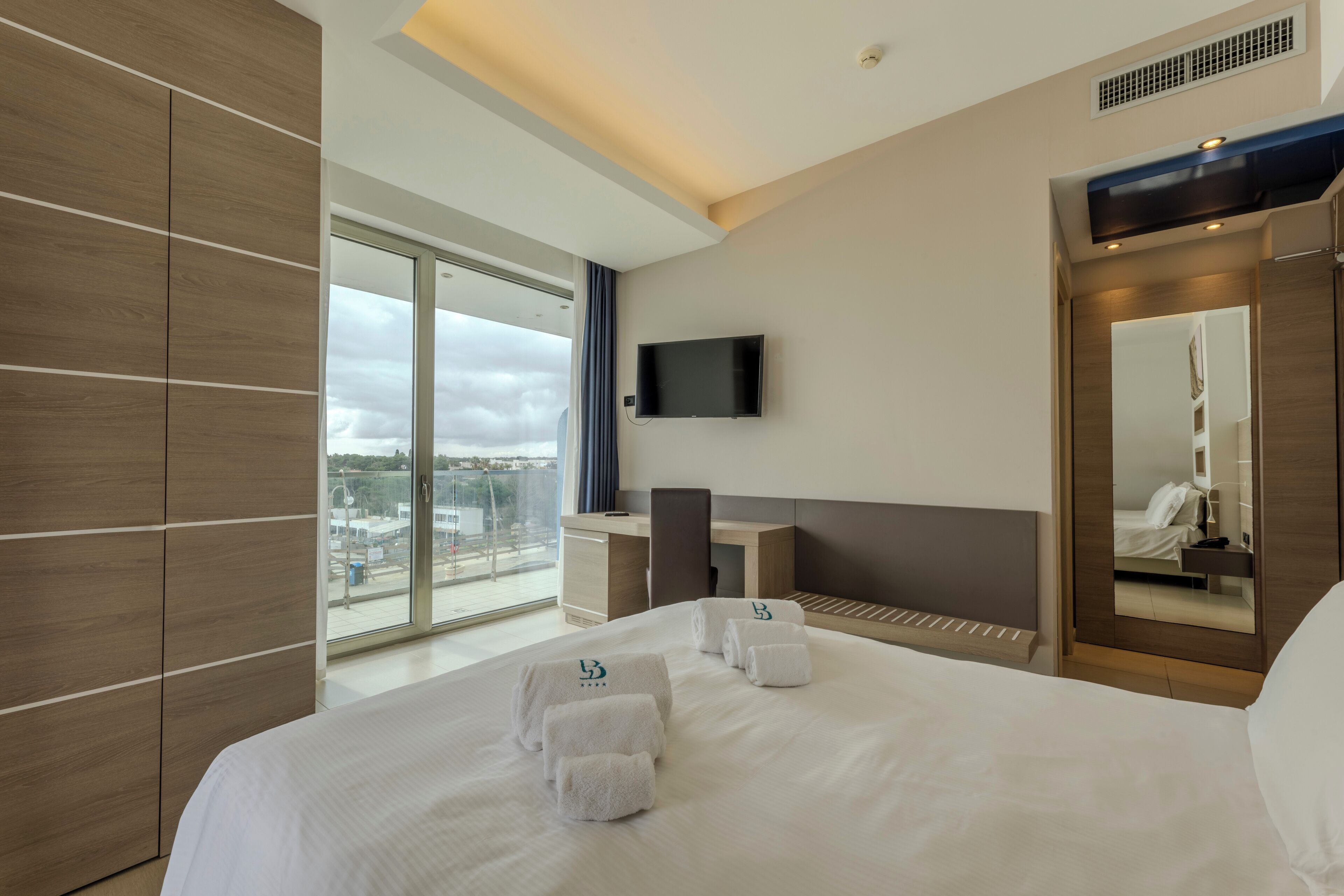A modern hotel room with a large bed, a desk, a chair, a TV, and a large window with a city view.