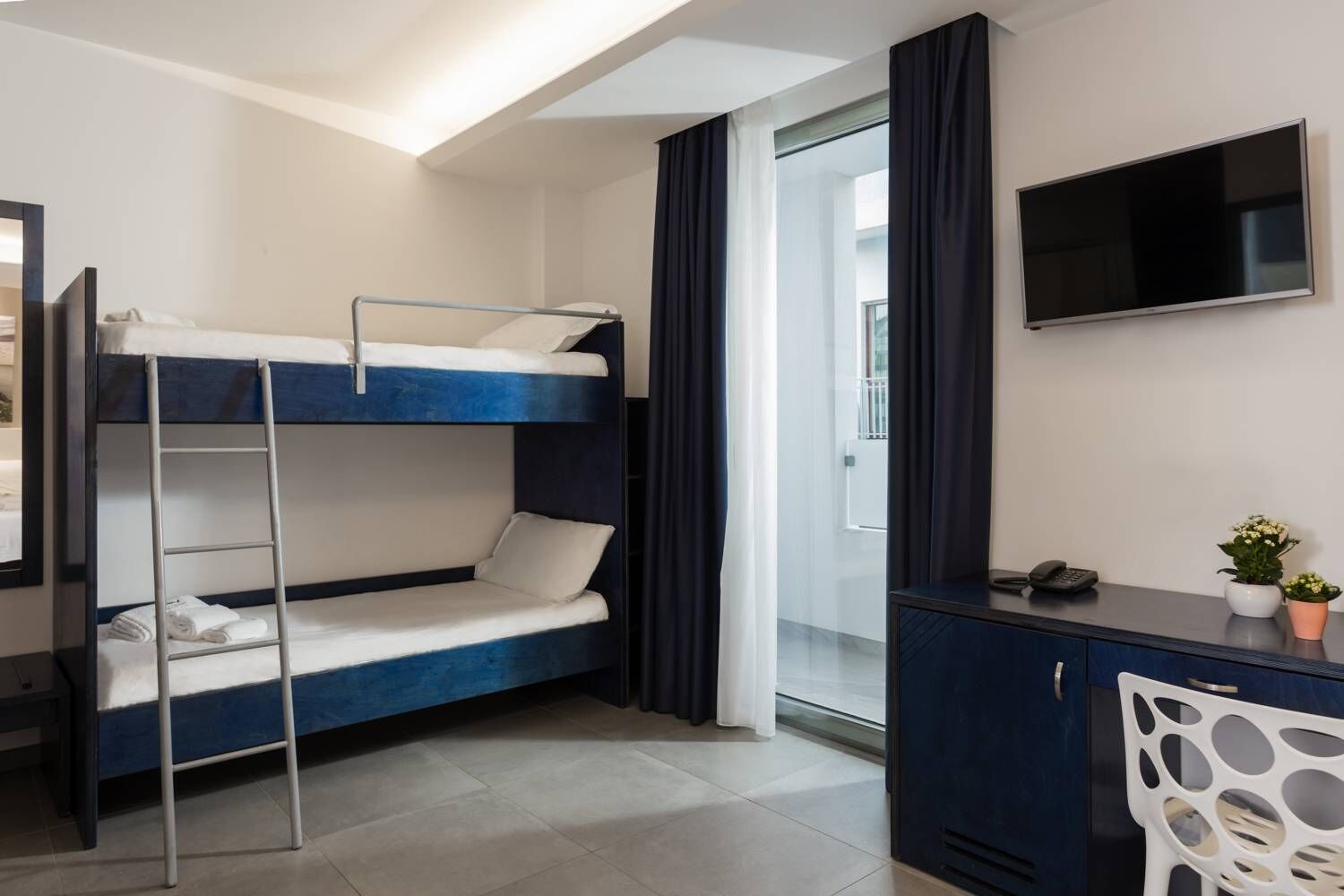 Cama individual superpuesta con escritorio, televisor, una silla y ventana con cortinas.