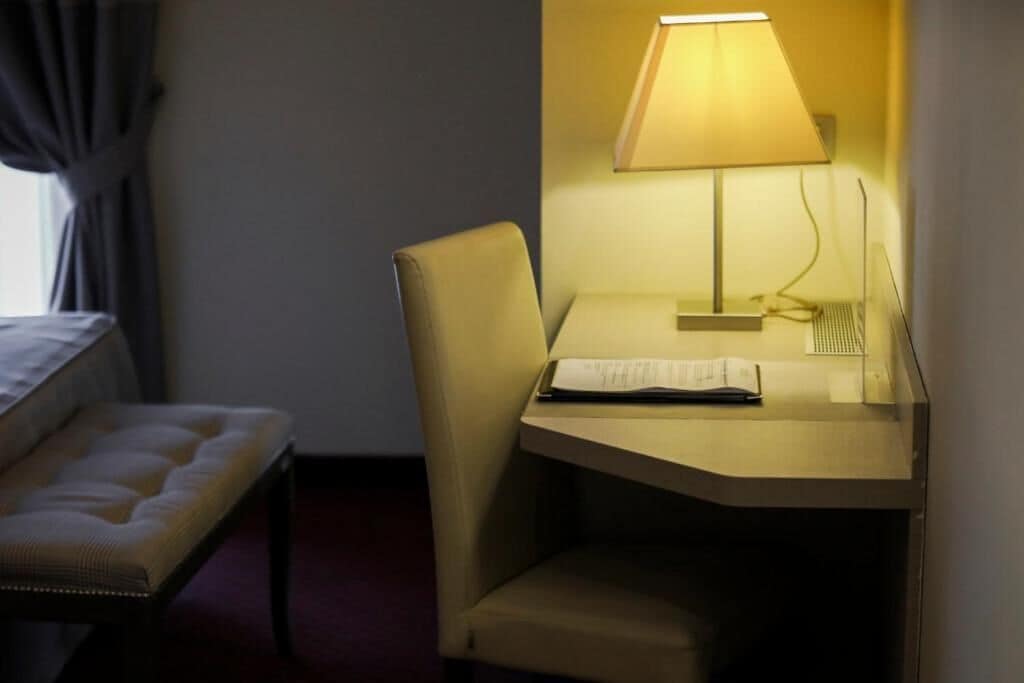 A hotel room with a bed, a chair, a desk with a lamp, and a window with curtains.