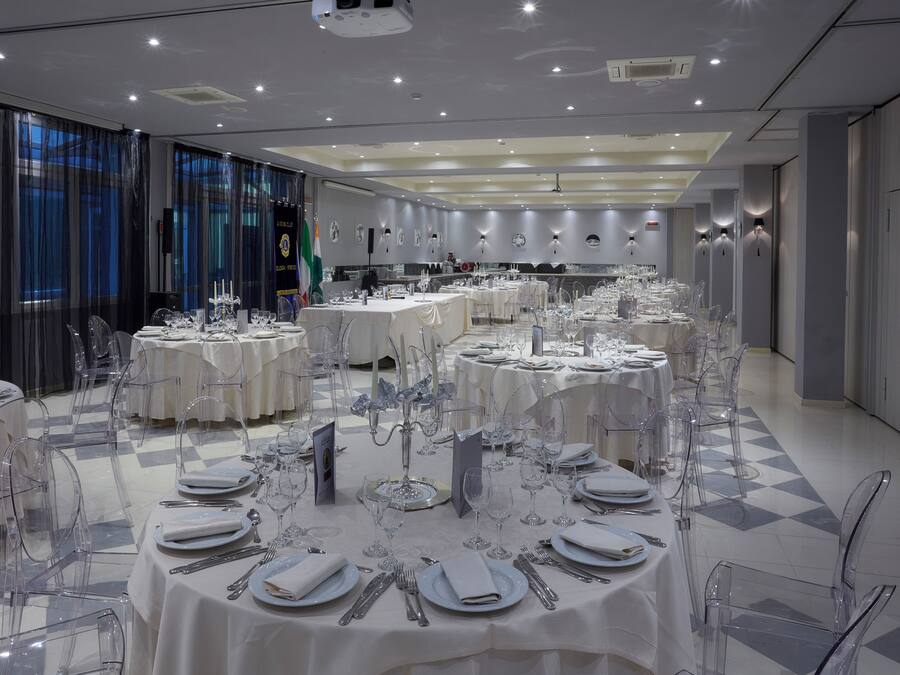 Un salón de banquetes con mesas redondas dispuestas para comer, sillas transparentes y piso a cuadros.