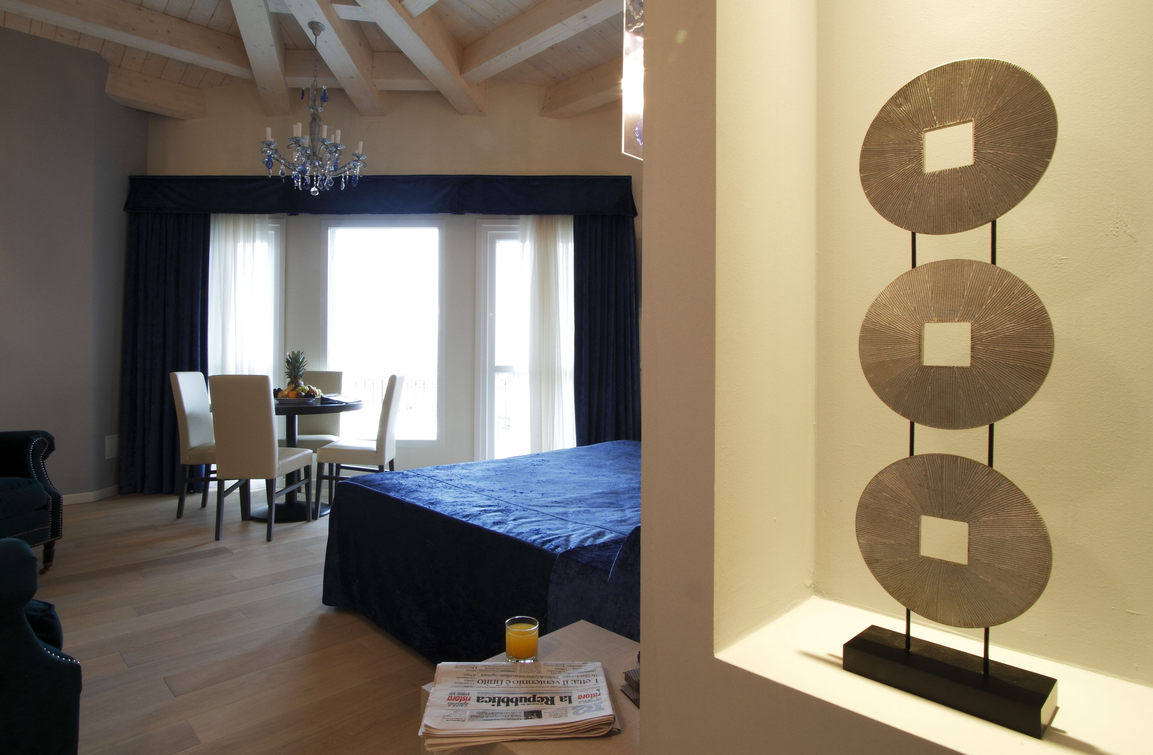 A hotel room with a bed, a dining table with chairs, a chandelier, and a decorative wall piece.