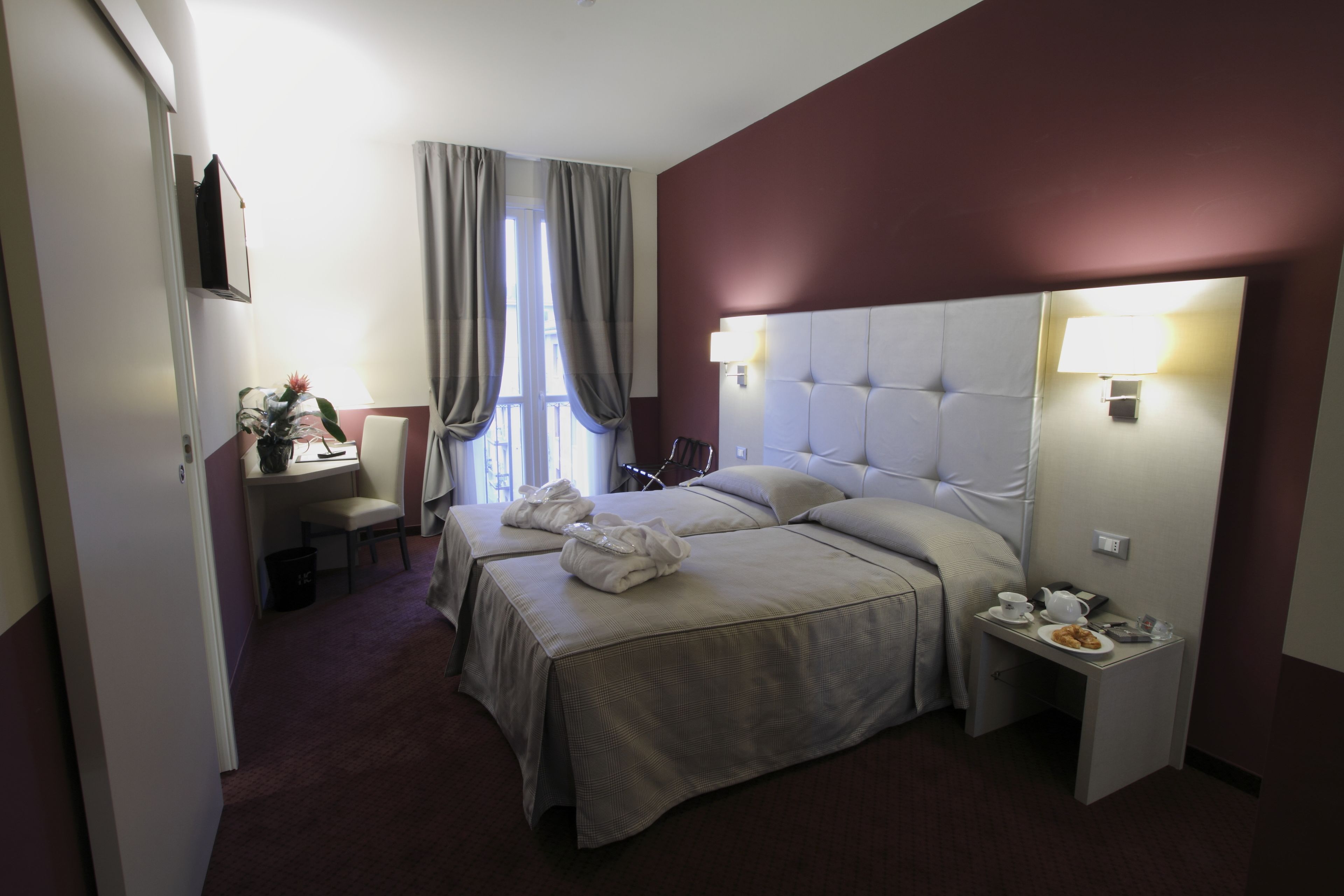 A hotel room with two beds, a desk with a chair, a small table with a lamp, and a window with curtains.