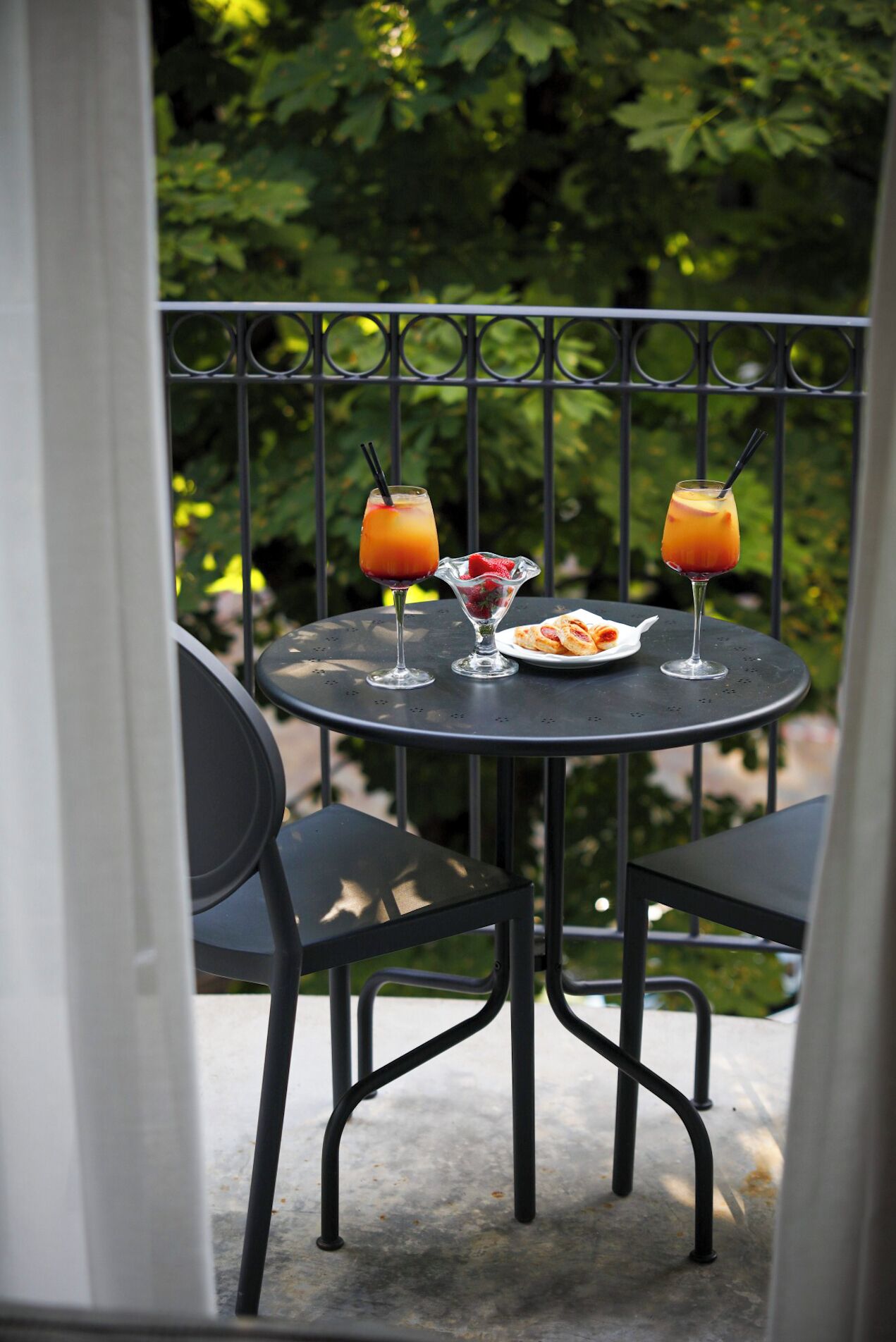 Une table ronde en métal, accompagnée de deux chaises, de deux boissons, d’une assiette de pâtisseries et d’un verre de fruits, est placée sur un balcon doté d’une rambarde en métal et dont l’arrière-plan est agrémenté de verdure.