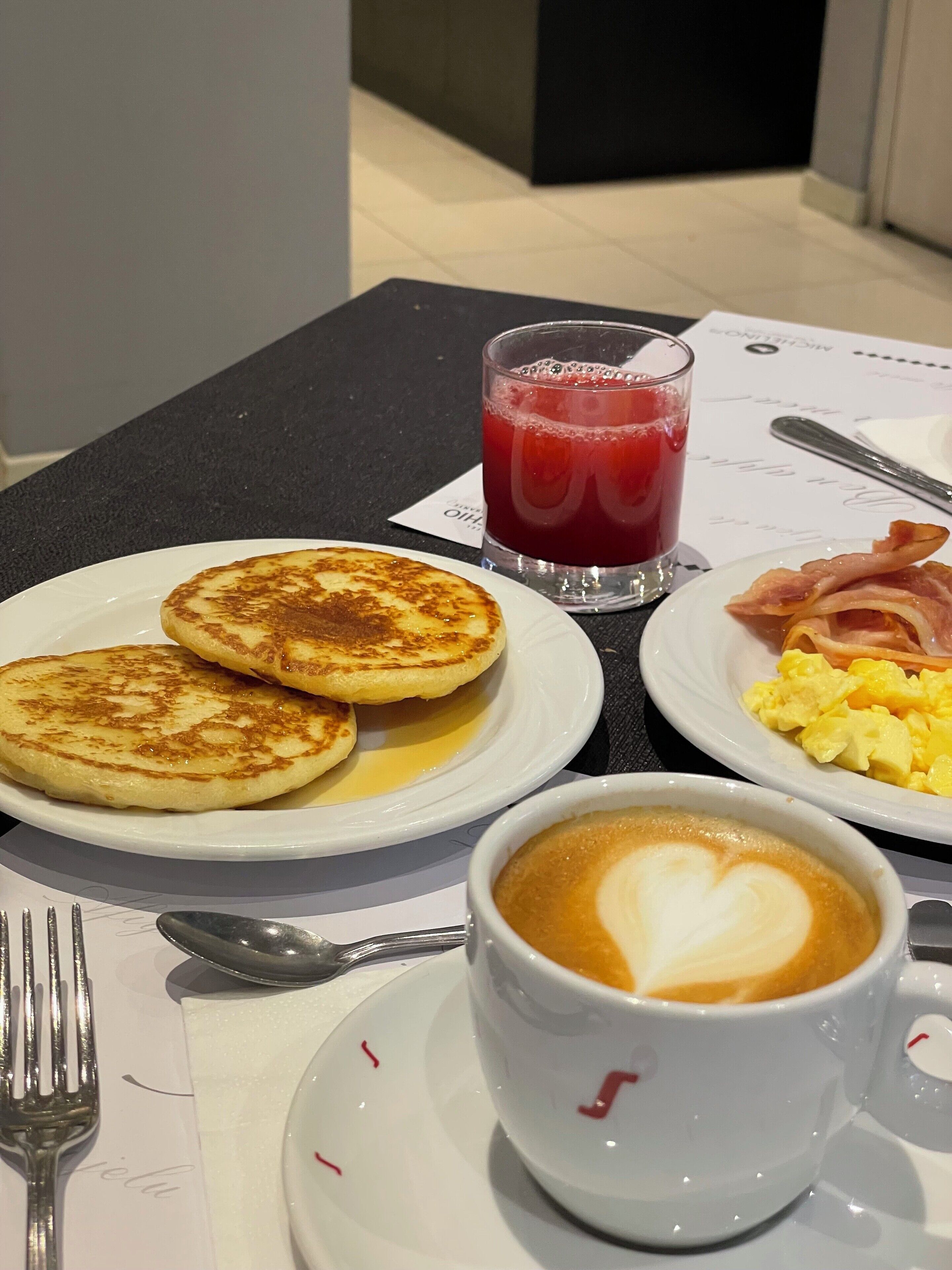 A breakfast table with pancakes, scrambled eggs, bacon, a glass of red juice, and a cup of coffee with latte art.