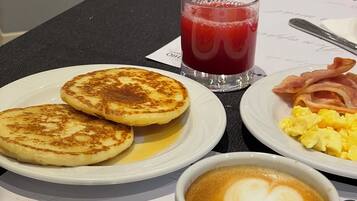 A breakfast table with pancakes, scrambled eggs, bacon, a glass of red juice, and a cup of coffee with latte art.