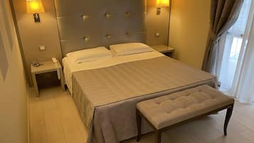 Ein Hotelzimmer mit einem großen Bett, zwei Nachttischen, einer Nachttischlampe, einer Bank und Blick ins Freie.