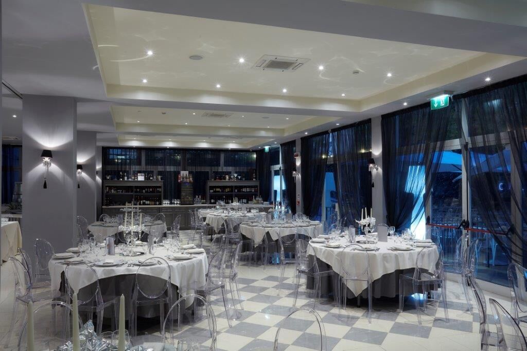 A restaurant with multiple round tables set for dining, clear chairs, and a checkered floor.