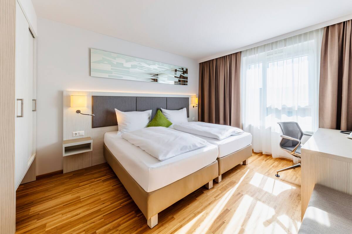 Hotellihuone, jossa on kaksi sänkyä, työpöytä ja suuri ikkuna, jossa on verhot.
