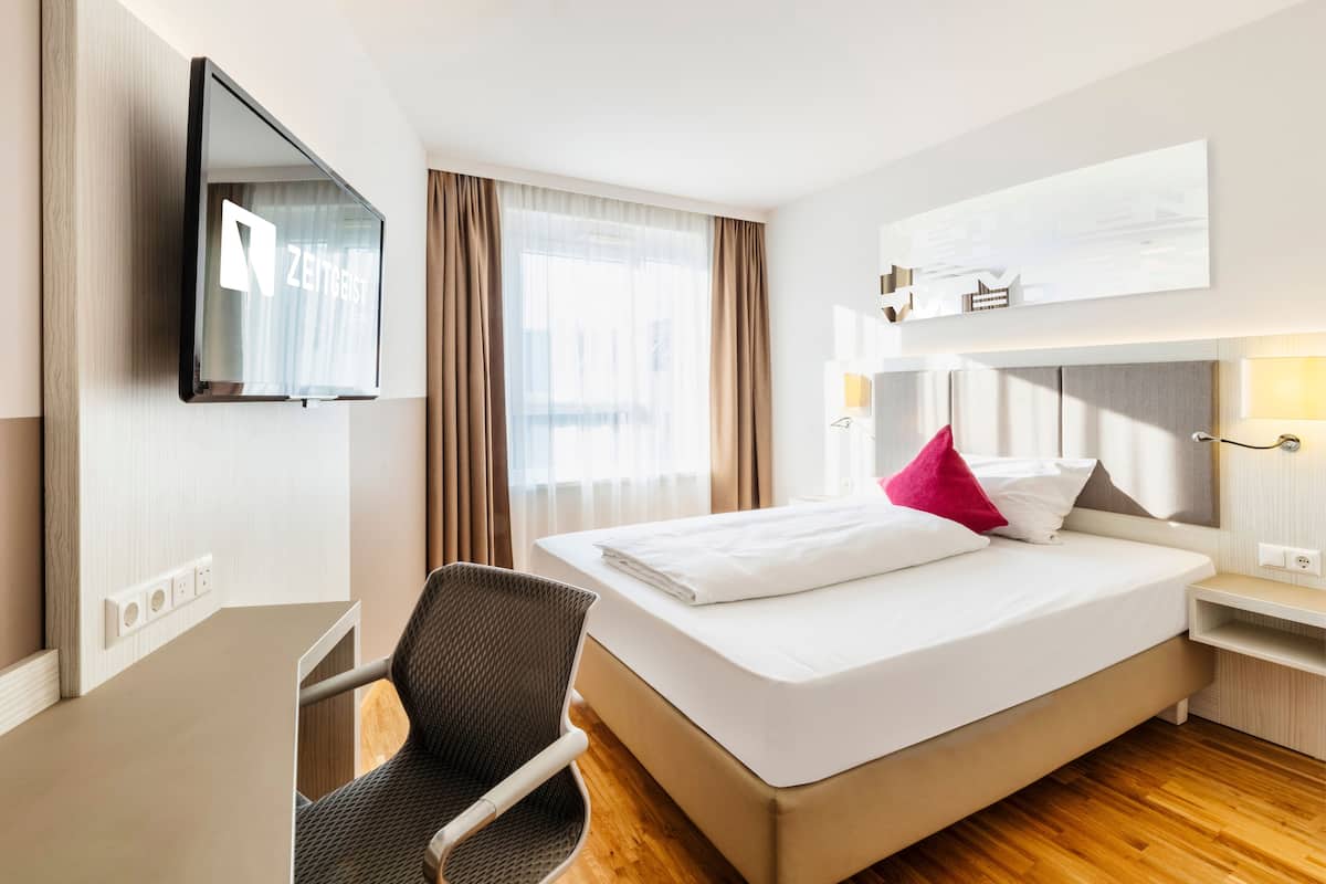 Hotellihuone, jossa on sänky, tuoli, seinälle asennettu televisio ja ikkuna, jossa on verhot.