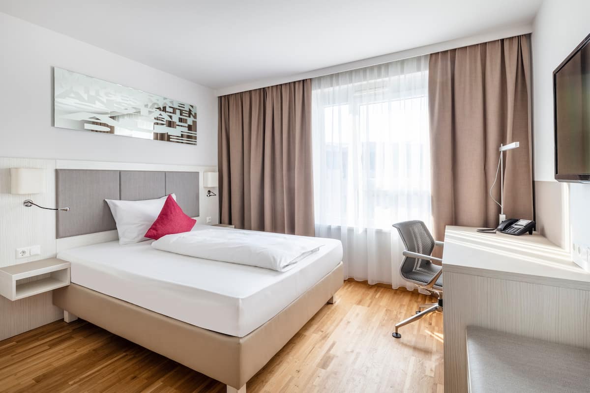 Hotellihuone, jossa on sänky, yöpöydät, työpöytä tuolilla, televisio ja ikkuna, jossa on verhot.