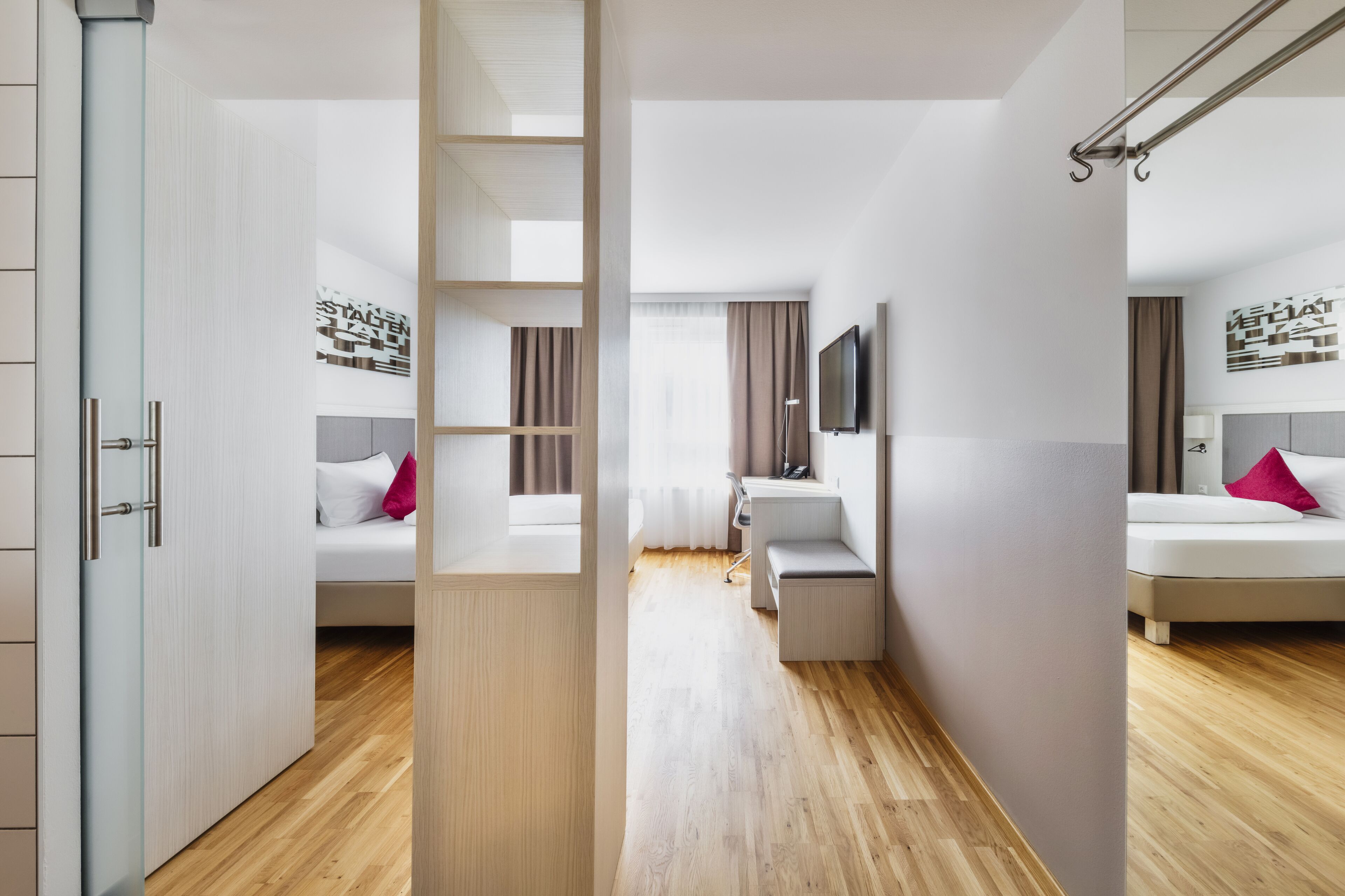 Quarto de hotel com piso de madeira, cama com cabeceira branca, um banco, televisão fixada na parede e uma janela com cortinas.