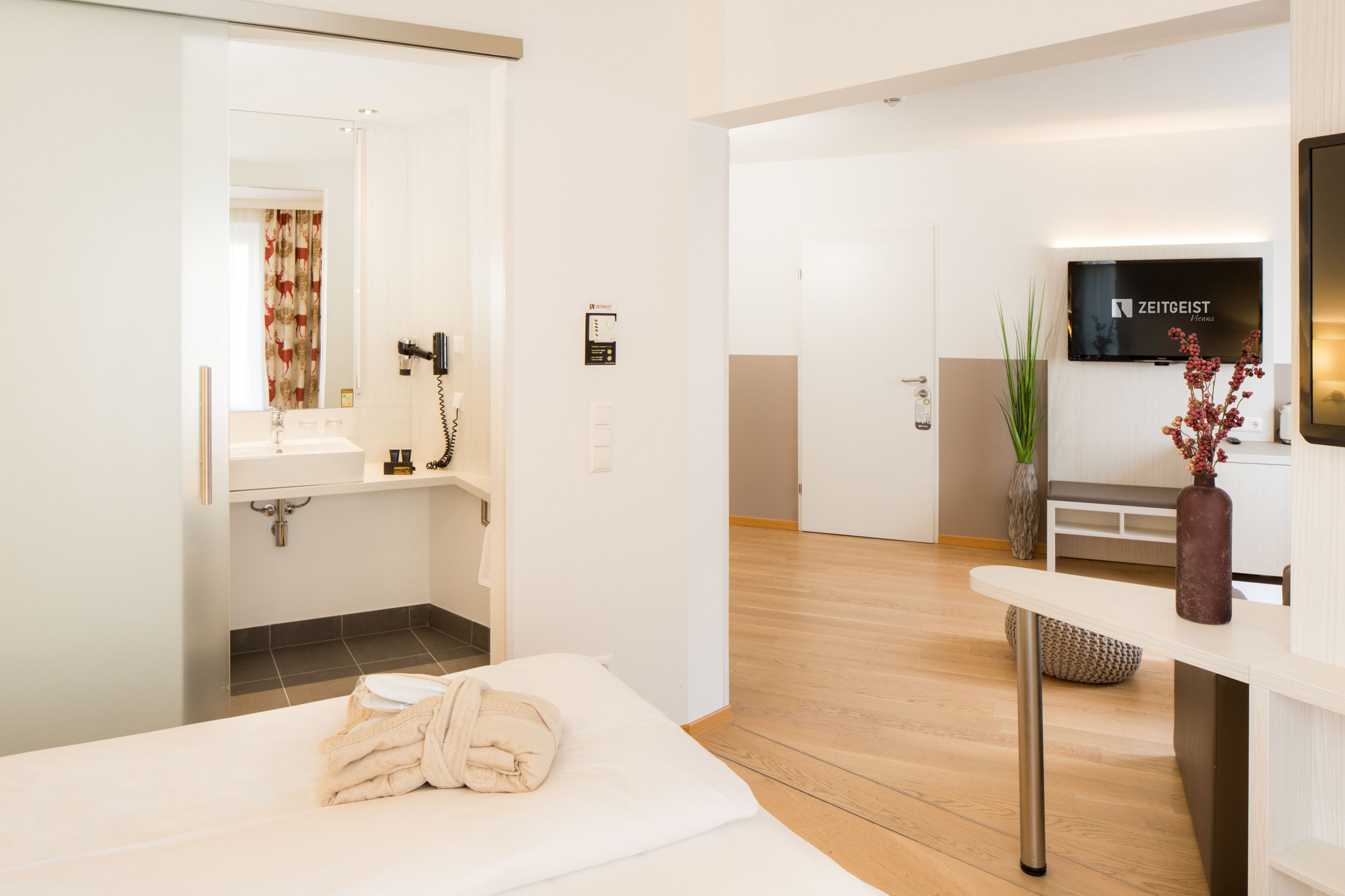 Une chambre d’hôtel moderne, dotée d’un lit, d’une salle de bain avec un lavabo et un miroir, d’un téléviseur fixé au mur et d’un plancher en bois.