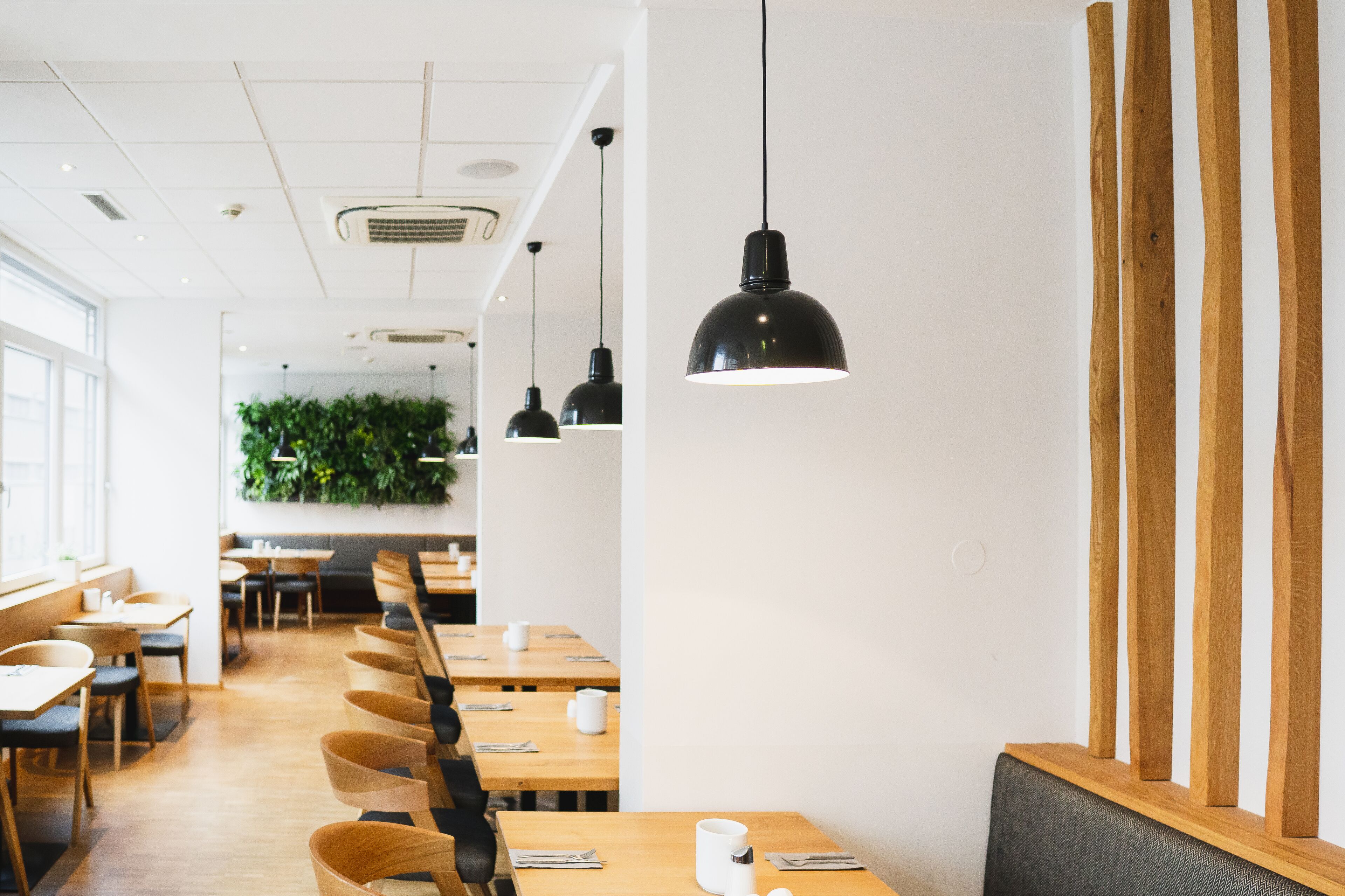 Un intérieur de restaurant moderne avec de longues tables en bois, des luminaires suspendus noirs et un mur végétal.