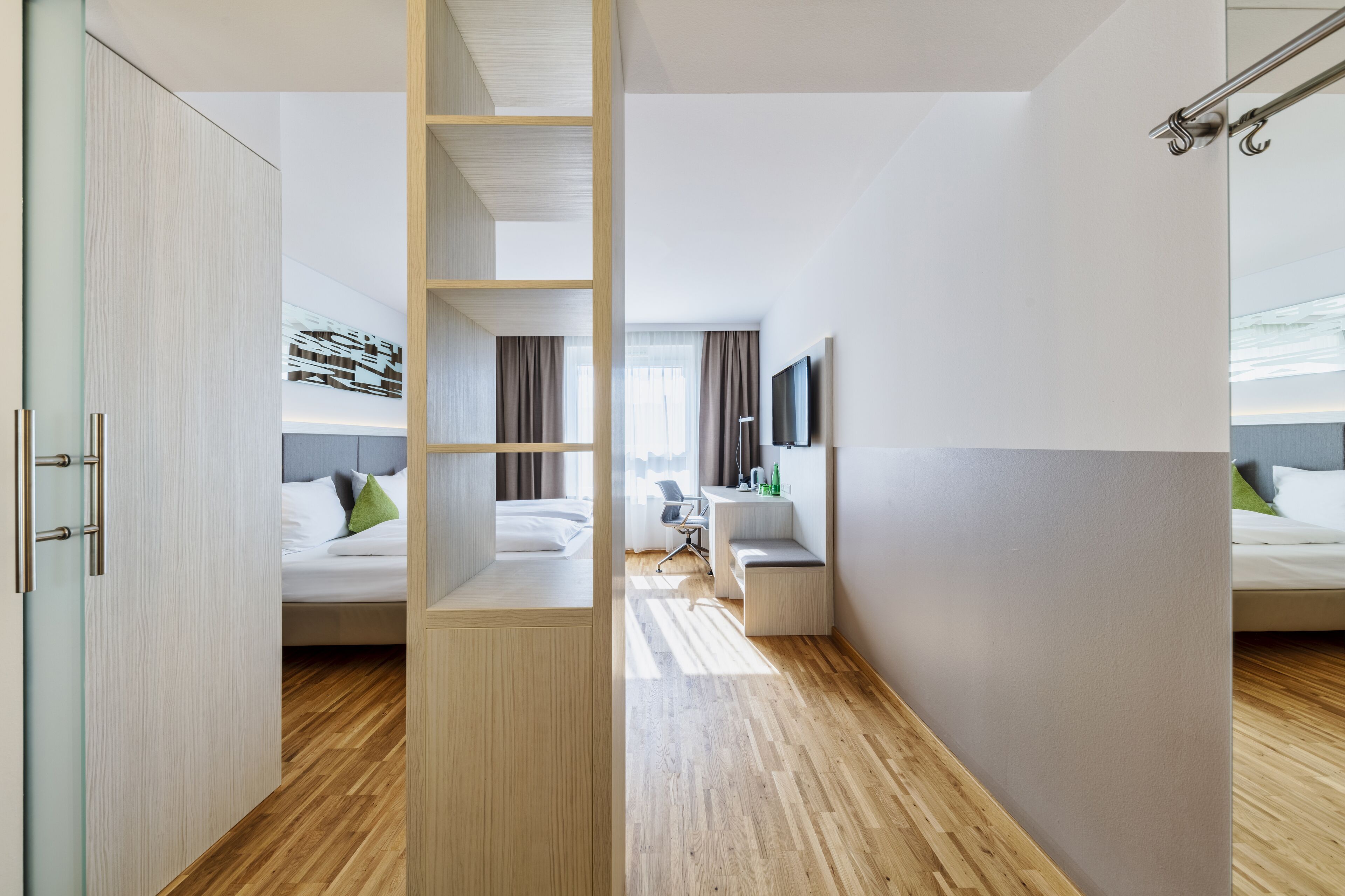 Quarto de hotel moderno com chão de madeira, cama com roupa de cama branca, escrivaninha com cadeira e televisão na parede.