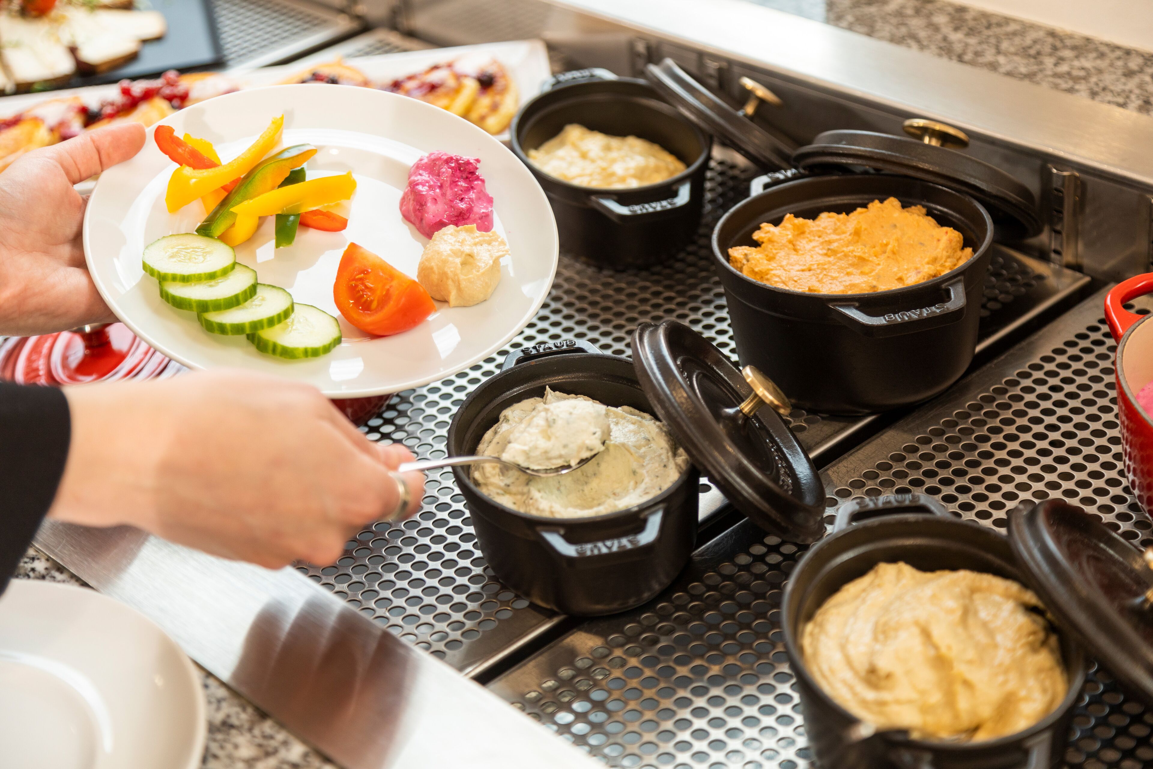 Uma pessoa se servindo em um buffet com diversos pratos, incluindo pepino fatiado, tomates e um prato com uma variedade de alimentos.