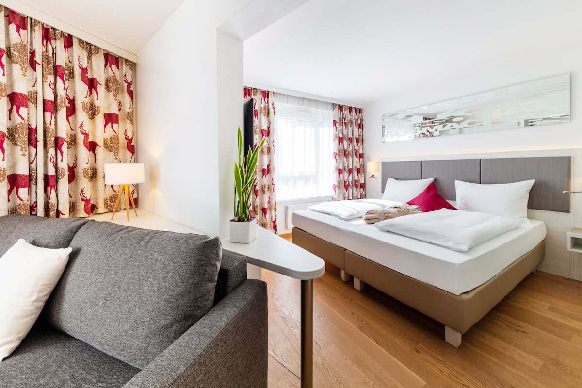 Moderni hotellihuone, jossa on sänky, harmaa sohva, pieni pöytä ja ruukussa oleva kasvi.