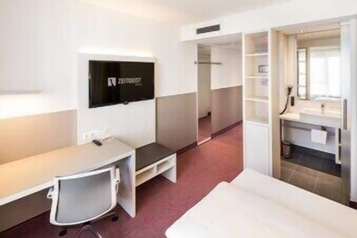 Hotellihuone, jossa on työpöytä, tuoli, televisio sekä kylpyhuone, jossa on pesuallas ja peili.