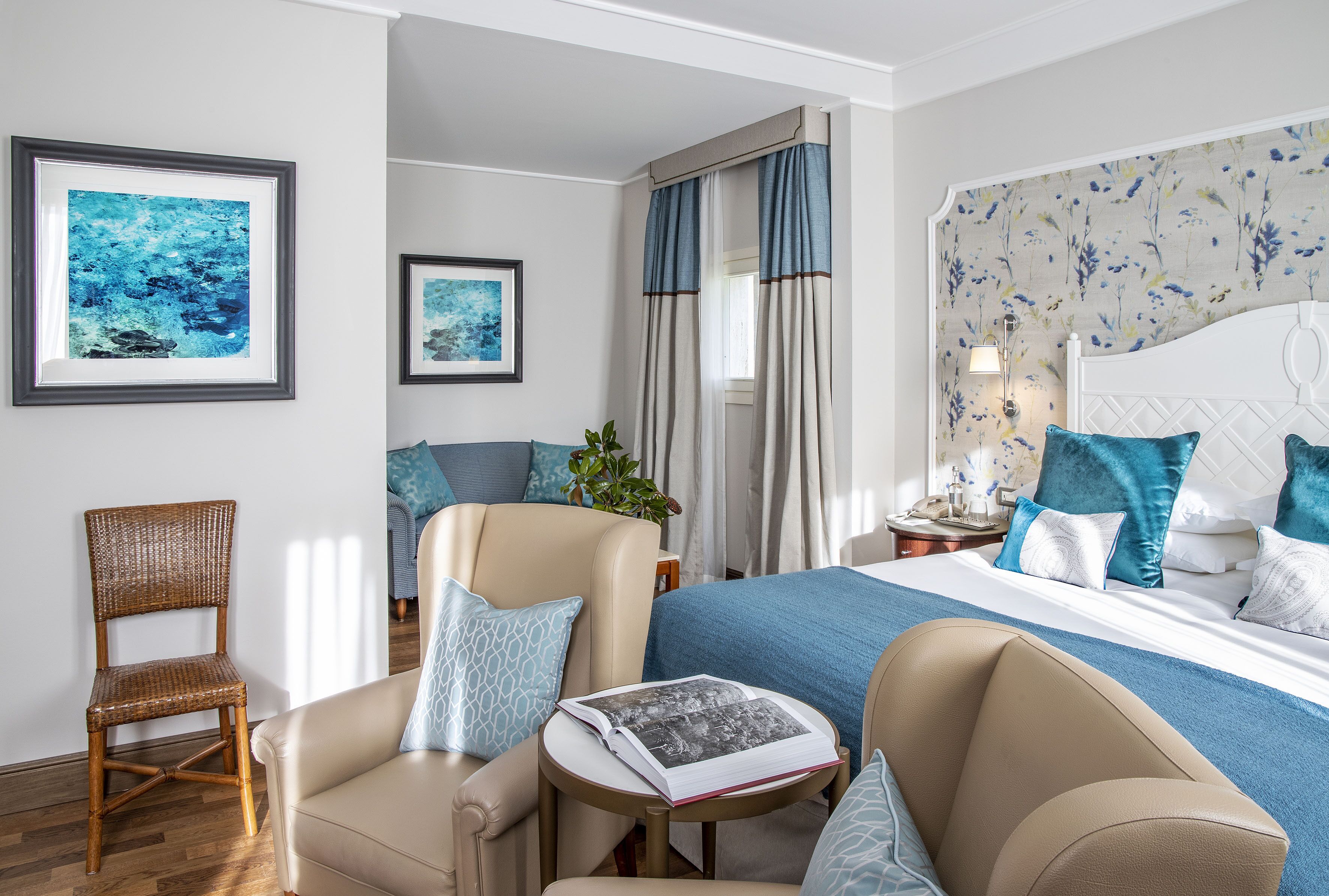 A hotel room with a bed, two armchairs, a small table with a book, and framed artwork on the wall.