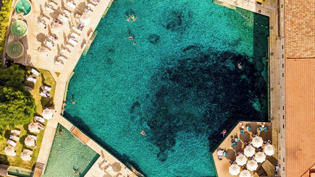 Vista aérea de una amplia piscina azul cristalina rodeada de tumbonas y sombrillas, con una zona de piscina más pequeña y un edificio contiguo a la piscina.