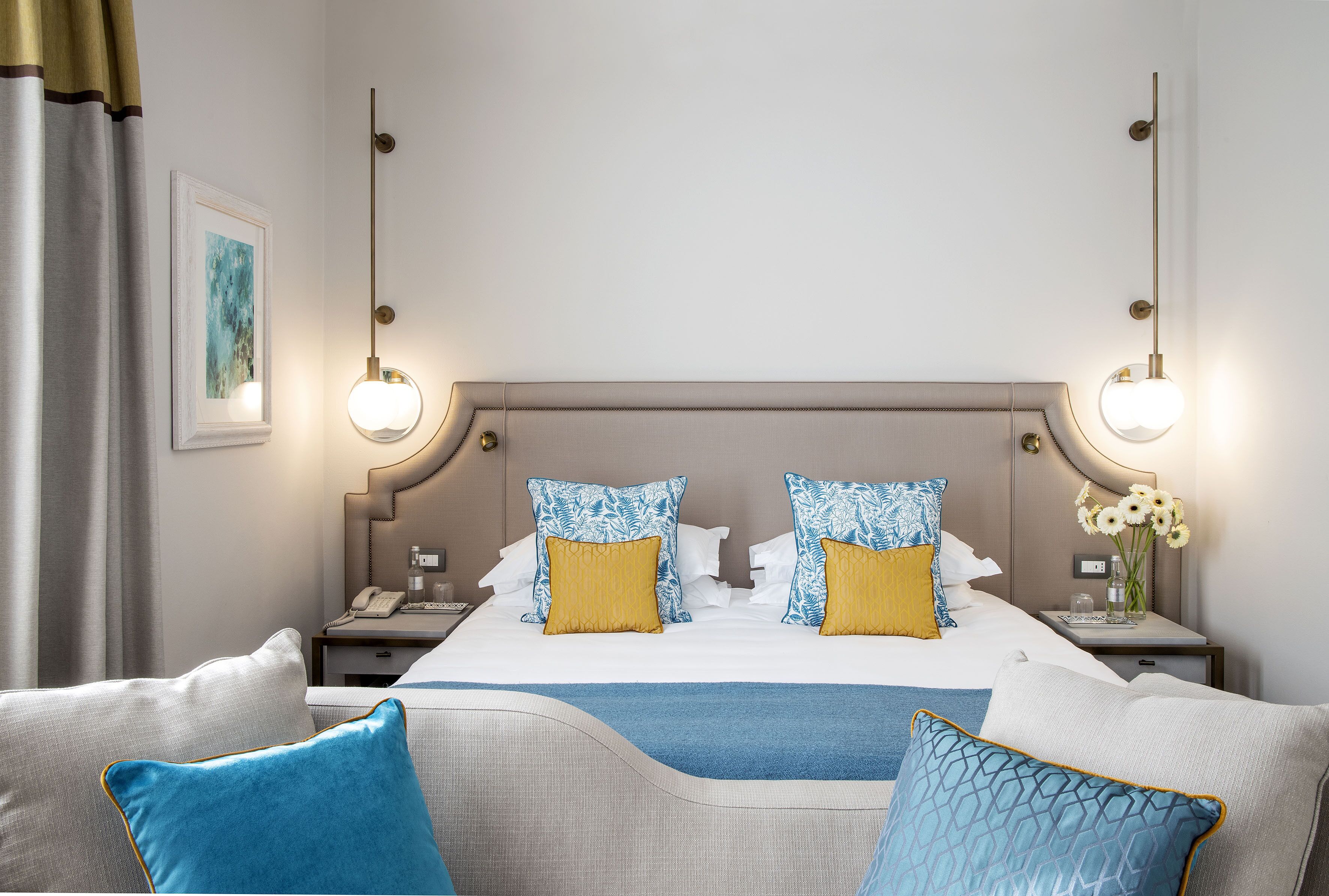 A hotel room with a large bed, two bedside tables, a nightstand, a wall-mounted lamp, and a framed picture.
