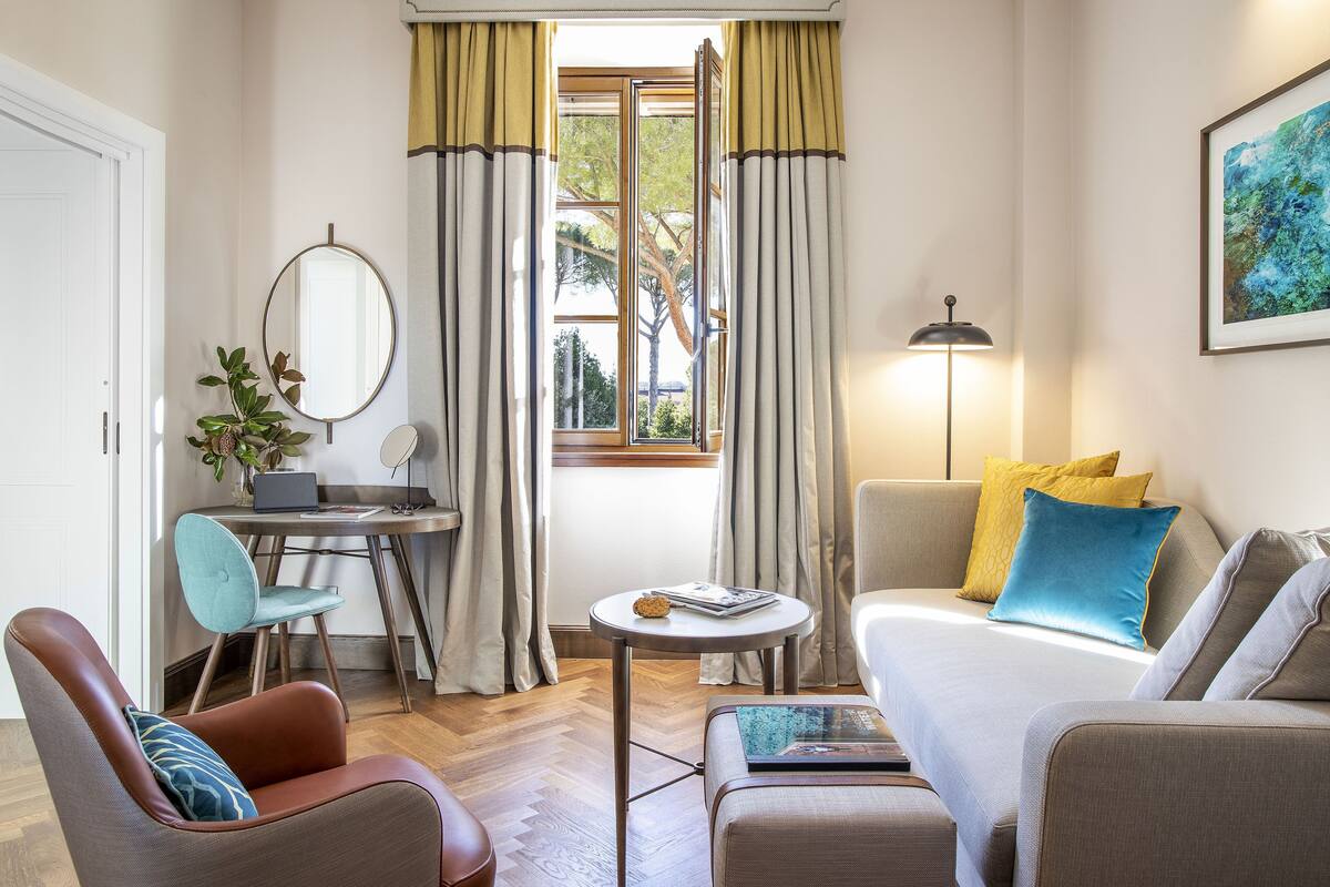 Ein kompakter Wohnbereich mit Sofa, Sessel, Schreibtisch und einem kleinen Tisch. Ein Fenster mit Vorhängen ist sichtbar, und eine Lampe steht rechts.