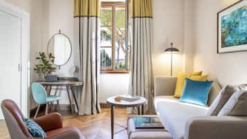 Um espaço de estar compacto com sofá, poltrona, escrivaninha e uma pequena mesa. Uma janela com cortinas é visível, e um abajur está à direita.