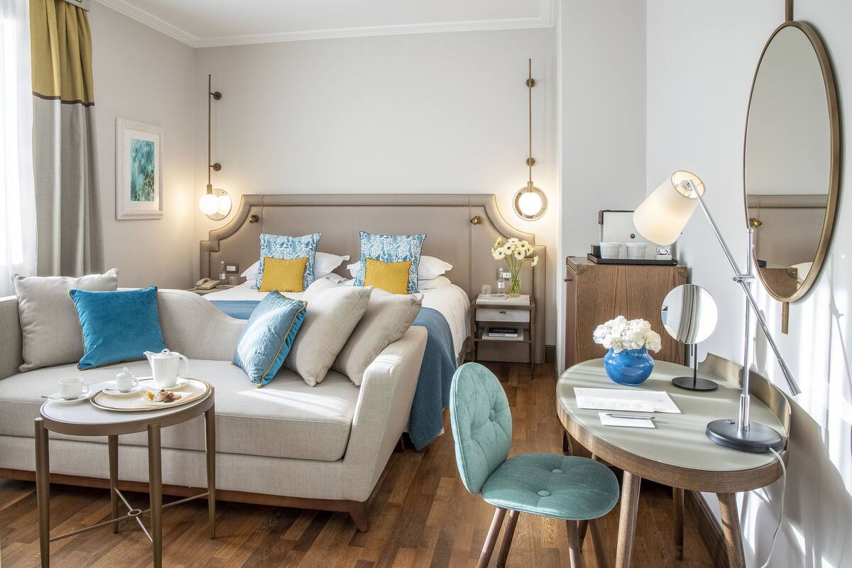 Ein Hotelzimmer mit Bett, Sofa, Esstisch mit Stühlen, Schreibtisch mit Stuhl, Spiegel und Blumen in einer Vase.