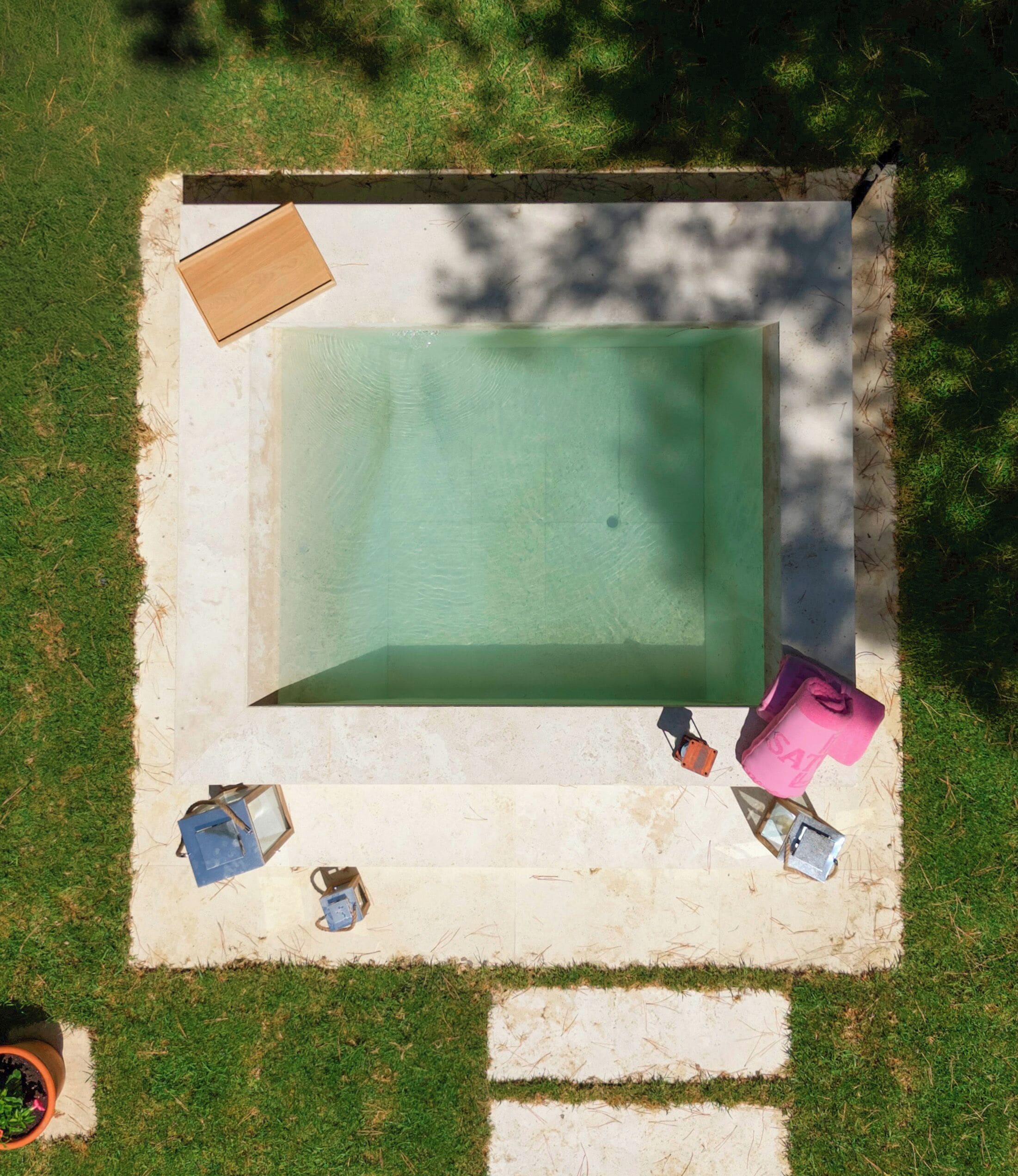 Una piscina rettangolare con una terrazza circostante, una panca in legno e alcune piante in vaso.