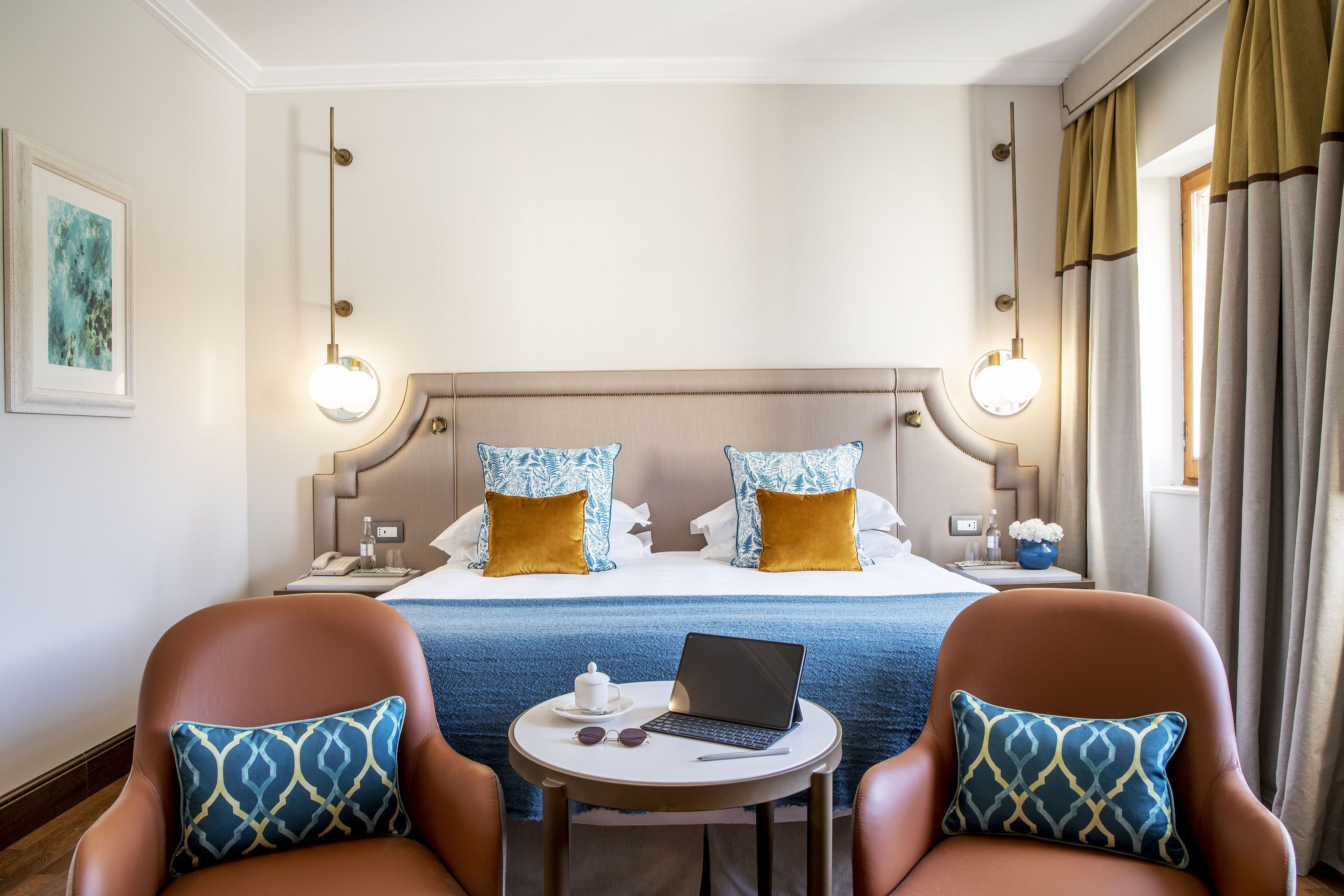 A hotel room with a bed, two armchairs, a small table with a laptop, and a bedside table with a lamp and a vase.